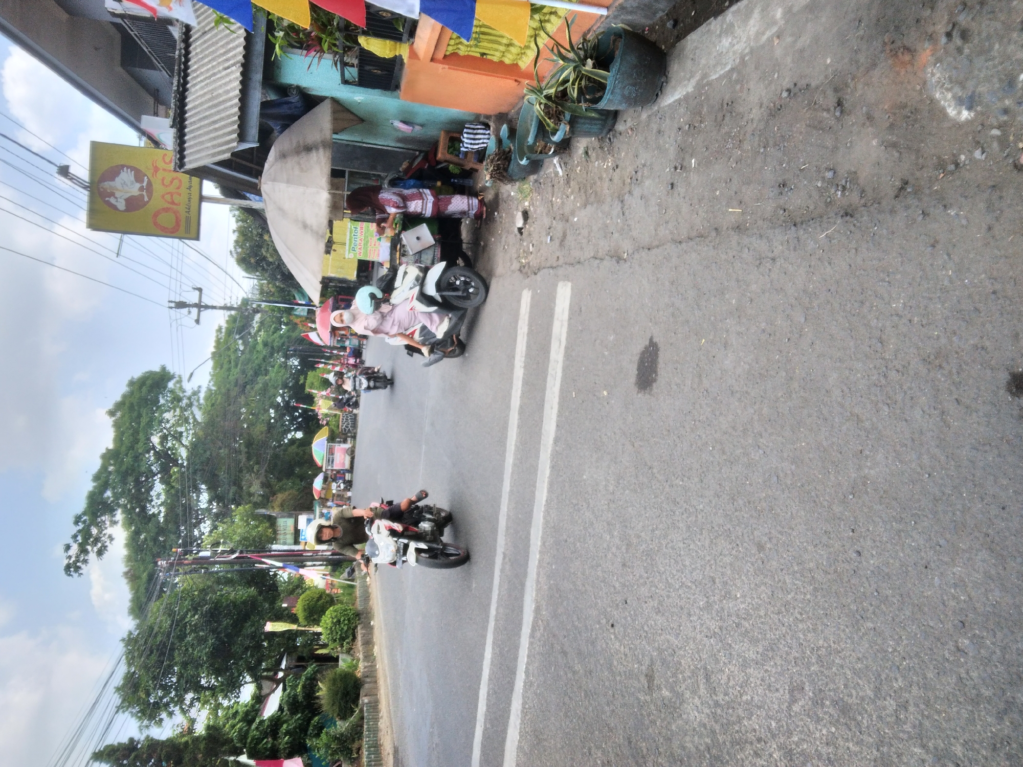 In the city of Sokaraja, Indonesia, a scene unfolds on a paved road with a sidewalk on the right. A person wearing a green shirt and a helmet rides a white and red motorcycle in the left lane. Further ahead, another motorcycle is visible in the right lane. On the sidewalk, a vendor under a beige umbrella is serving a customer seated on a white scooter. The vendor wears a patterned maroon and white outfit, while the customer is dressed in pink and a light blue face mask. Behind them, a yellow sign with a chicken illustration and the word "Oasis" is mounted on a building. The environment includes trees, power lines, and various potted plants on the sidewalk. The sky is cloudy.