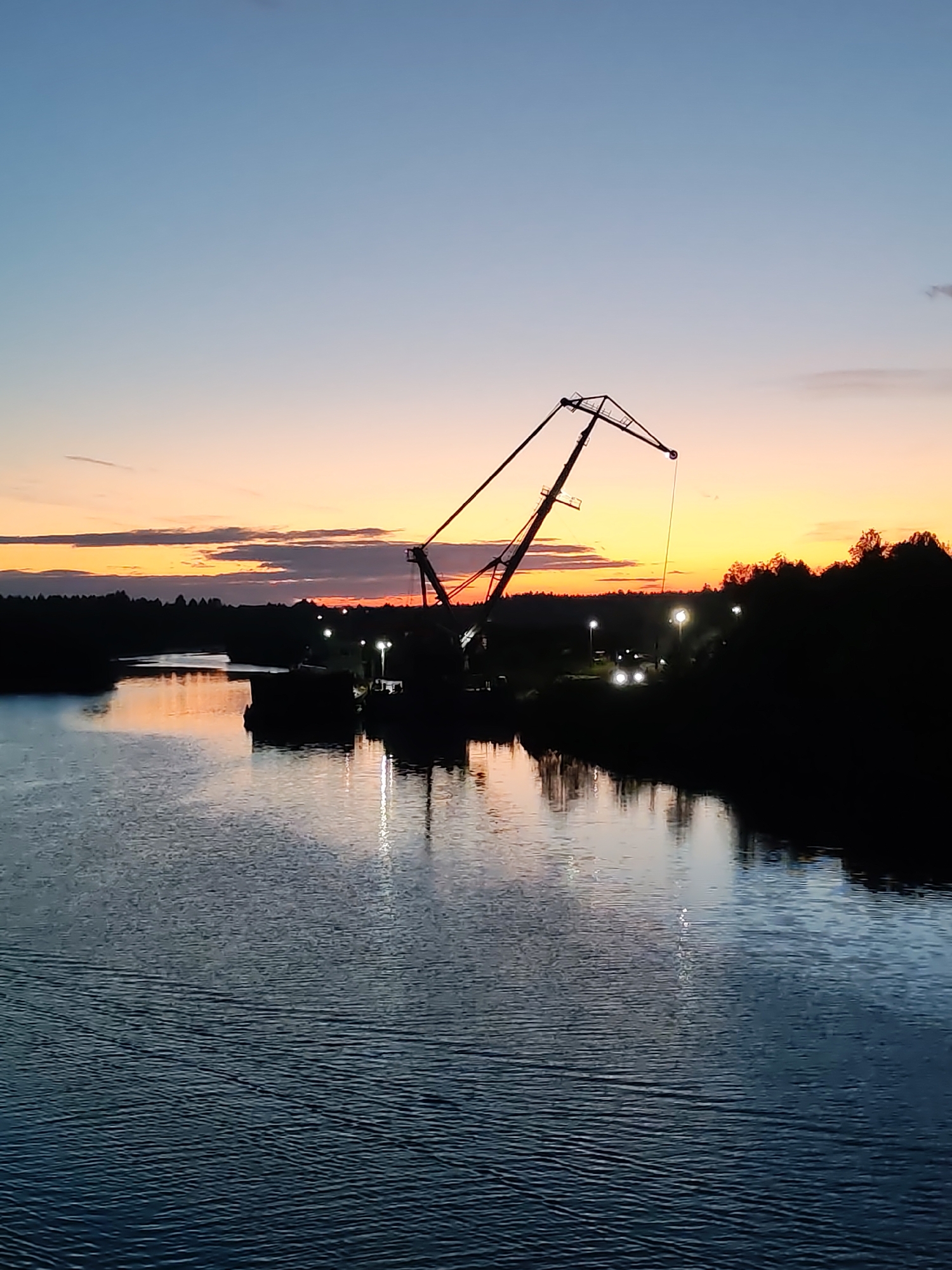 The image depicts a river scene at dusk in Annensky Most, Russia. A large crane is silhouetted against the colorful sunset sky, positioned on a barge or dock along the riverbank. The water reflects the colors of the sky and the lights from the shore. The opposite bank is lined with dark trees and vegetation. Several lights are visible along the shoreline. The overall scene is one of industrial activity against a natural backdrop during twilight.