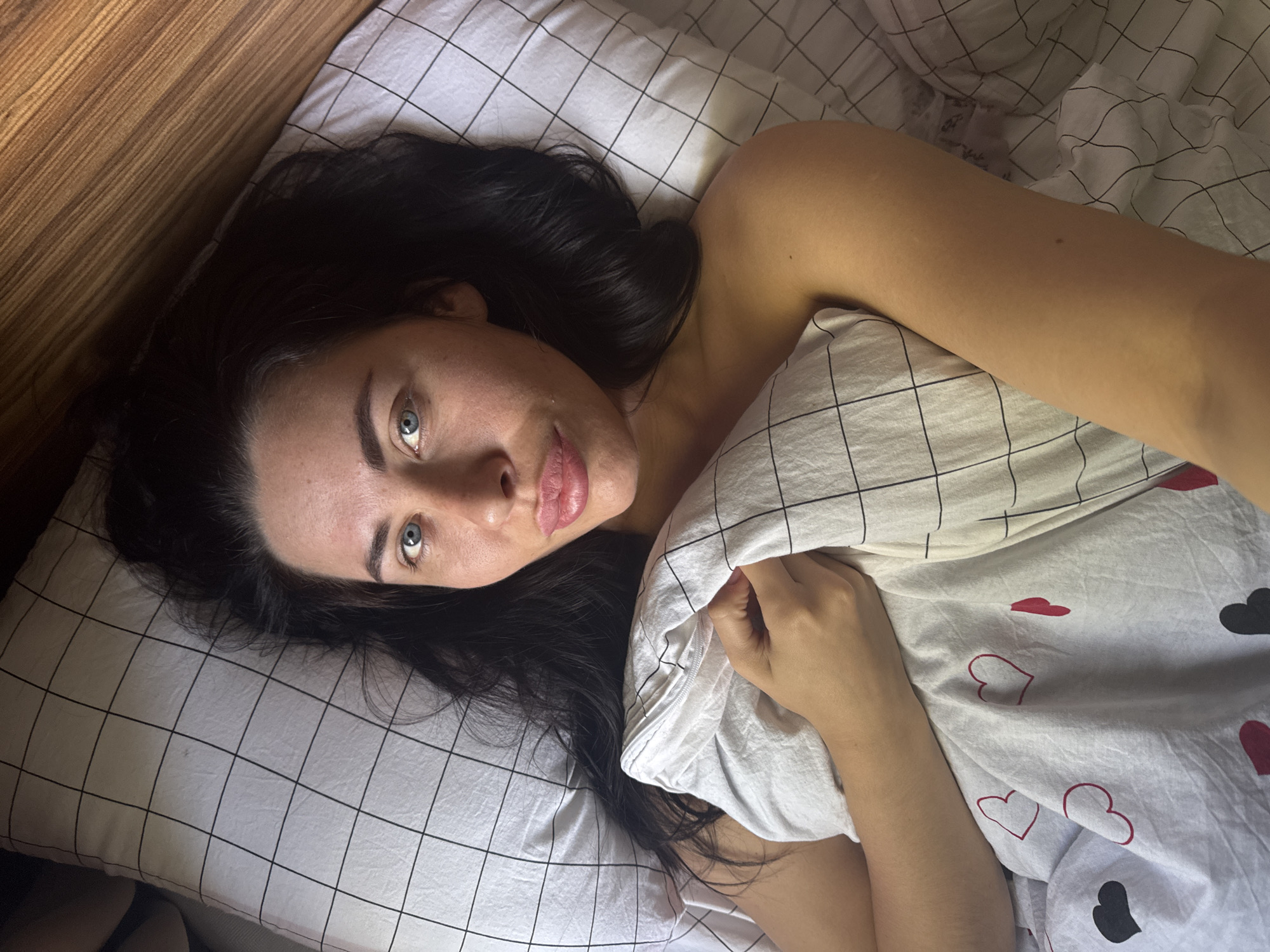 A young woman is captured in a medium-shot, slightly overhead photograph, lying in bed and looking directly at the camera. She has long, dark hair splayed across a white pillow adorned with a black grid pattern. Her prominent blue eyes are open, and her lips are softly parted.
Her upper body is partially visible, with her bare right shoulder exposed, resting on the grid-patterned bedding. Her right arm is bent, holding a white duvet that covers the lower part of her body. The duvet displays two distinct patterns: the section near her face features the same black grid as the pillow, while the portion covering her torso is decorated with a scattered design of red and black hearts, some solid and others outlined.
To the left of the woman, a textured wooden surface, possibly a headboard or wall panel, runs diagonally across the frame. The overall lighting is soft and diffused, indicating an indoor setting, likely a bedroom. No specific time of day or weather conditions can be inferred. The woman's expression is neutral and direct. There is no visible text in the image. The setting is contextually provided as Chaikovsky, Russia.