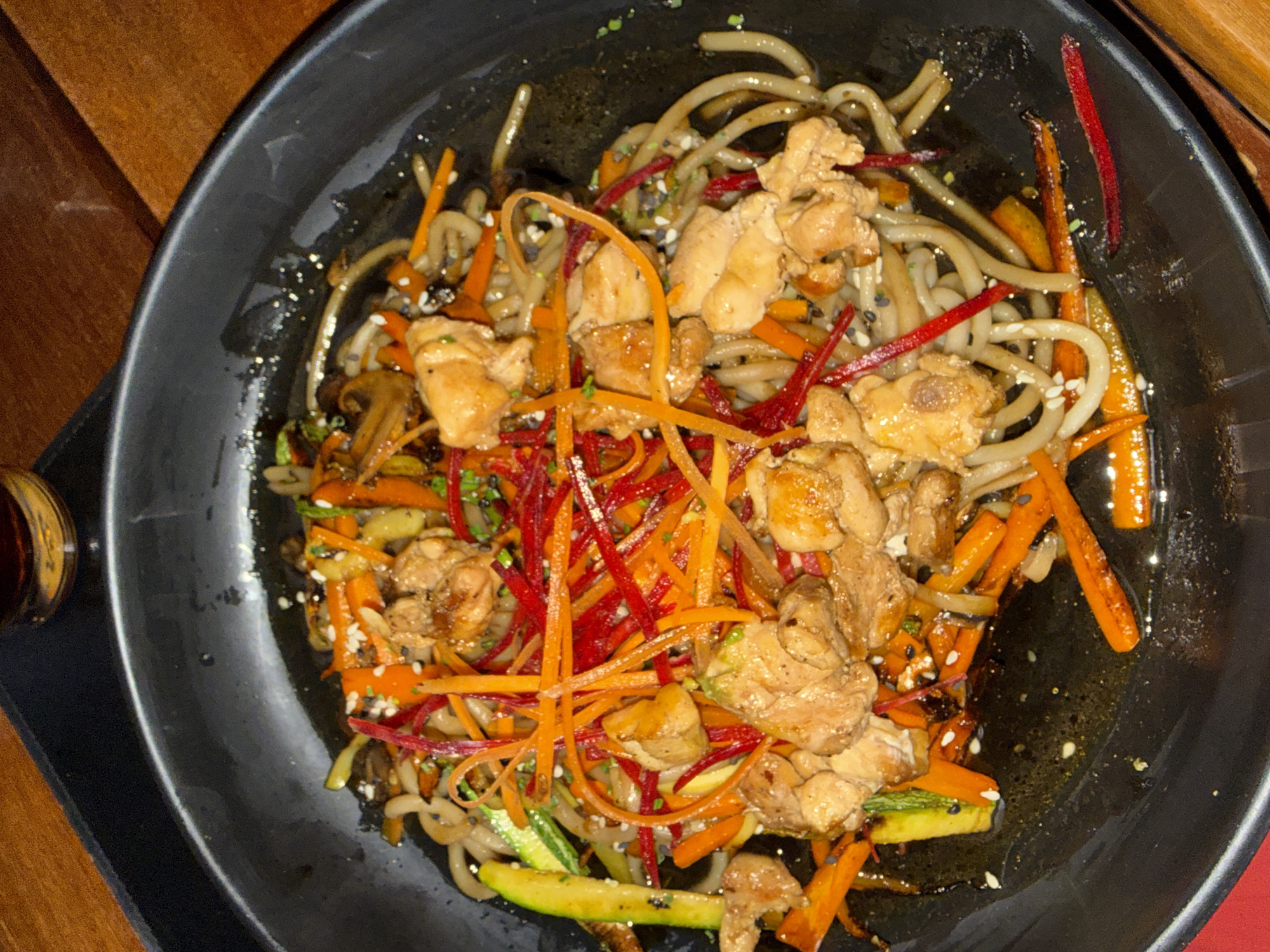 A top-down, close-up shot shows a black bowl filled with stir-fried noodles, chicken, and vegetables. The noodles are light gray or brown. Diced chicken pieces are scattered throughout the dish. Shredded carrots and beets are present, as well as zucchini. Sesame seeds are sprinkled across the dish. The bowl sits on a dark-colored placemat on a wooden table. A small container of sauce is visible on the left side of the frame. The photograph was taken in San Bernardino Tlaxcalancingo, Mexico.