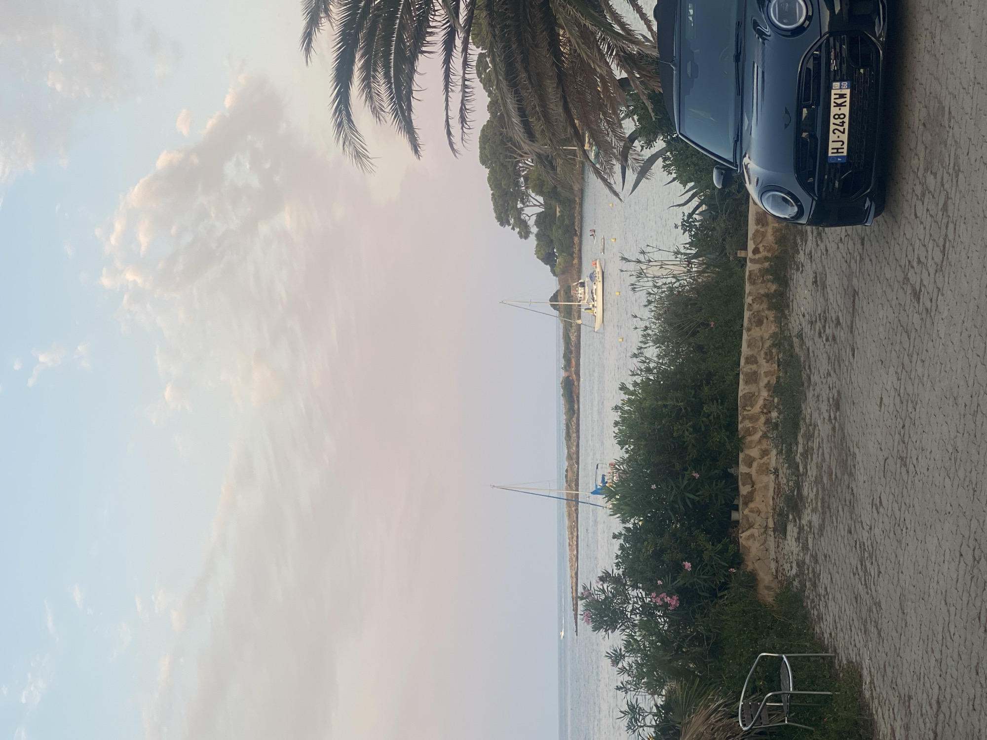 The image depicts a seaside scene in Antibes, France, with a black convertible Mini Cooper parked in the foreground on a paved driveway. The car's license plate reads "HJ-248-KW". To the left of the car, a silver metal chair sits on a patch of grass. Behind the chair and car, a low stone wall separates the paved area from lush green vegetation, including flowering bushes with pink blossoms. Beyond the greenery, the calm sea stretches out to the horizon. A white sailboat with blue accents is anchored in the water, and a small white boat with an orange stripe is visible further out at sea. In the distance, a tree-covered peninsula juts into the water, and palm fronds are visible in the upper right corner of the frame. The sky is a soft gradient of pale blue and pink, with scattered clouds, suggesting either sunrise or sunset.