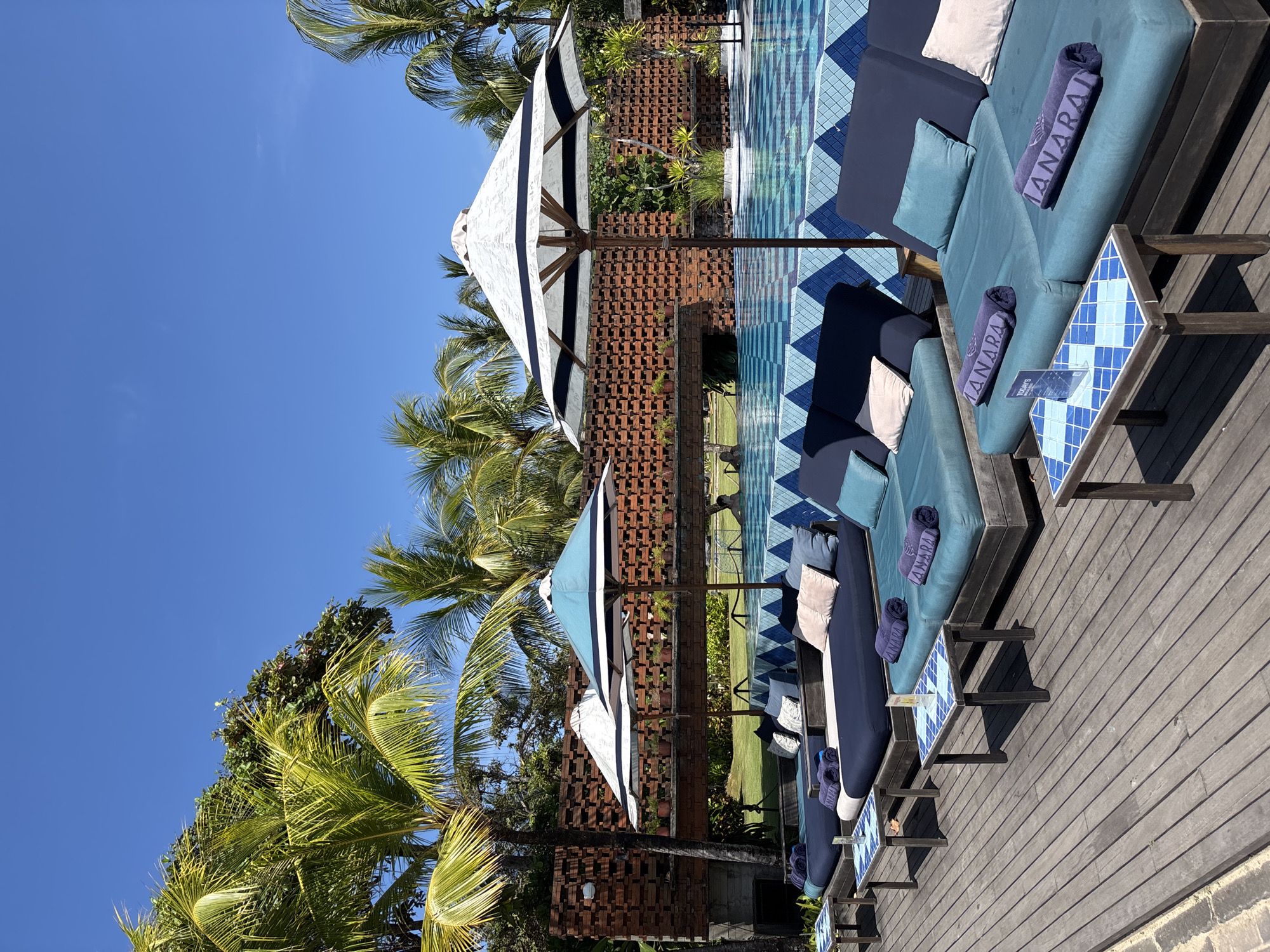 The image depicts a luxury poolside scene, likely at a resort, in Kuta Selatan, Indonesia. Rows of blue lounge chairs with dark blue accents and plush cushions are arranged along a wooden deck. Each lounger is adorned with a rolled purple towel labeled "ANARAJA" and a small, blue-tiled side table.
White and blue striped umbrellas provide shade. The pool adjacent to the loungers has a geometric, blue-tiled bottom. A brick wall decorated with potted plants acts as a backdrop, while palm trees are visible against a clear blue sky. There are no people in the frame.