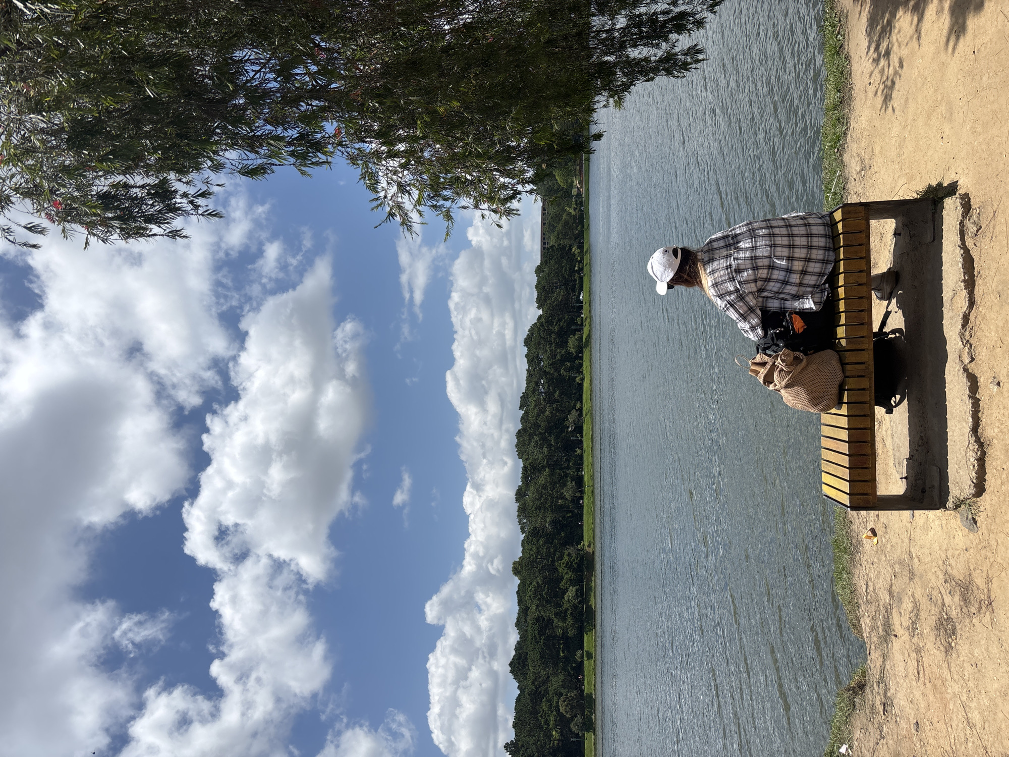 A person wearing a white baseball cap and a plaid shirt sits on a yellow bench by a lake. The person has their back to the camera, looking out at the water. A straw-colored bag and a black backpack are placed on the bench next to them. The lake is calm, with trees lining the opposite shore. The sky is blue with scattered white clouds. The scene appears to be in a park or natural area. This location is likely Xuan Huong Lake in Da Lat, Vietnam, known for its scenic beauty and recreational activities.