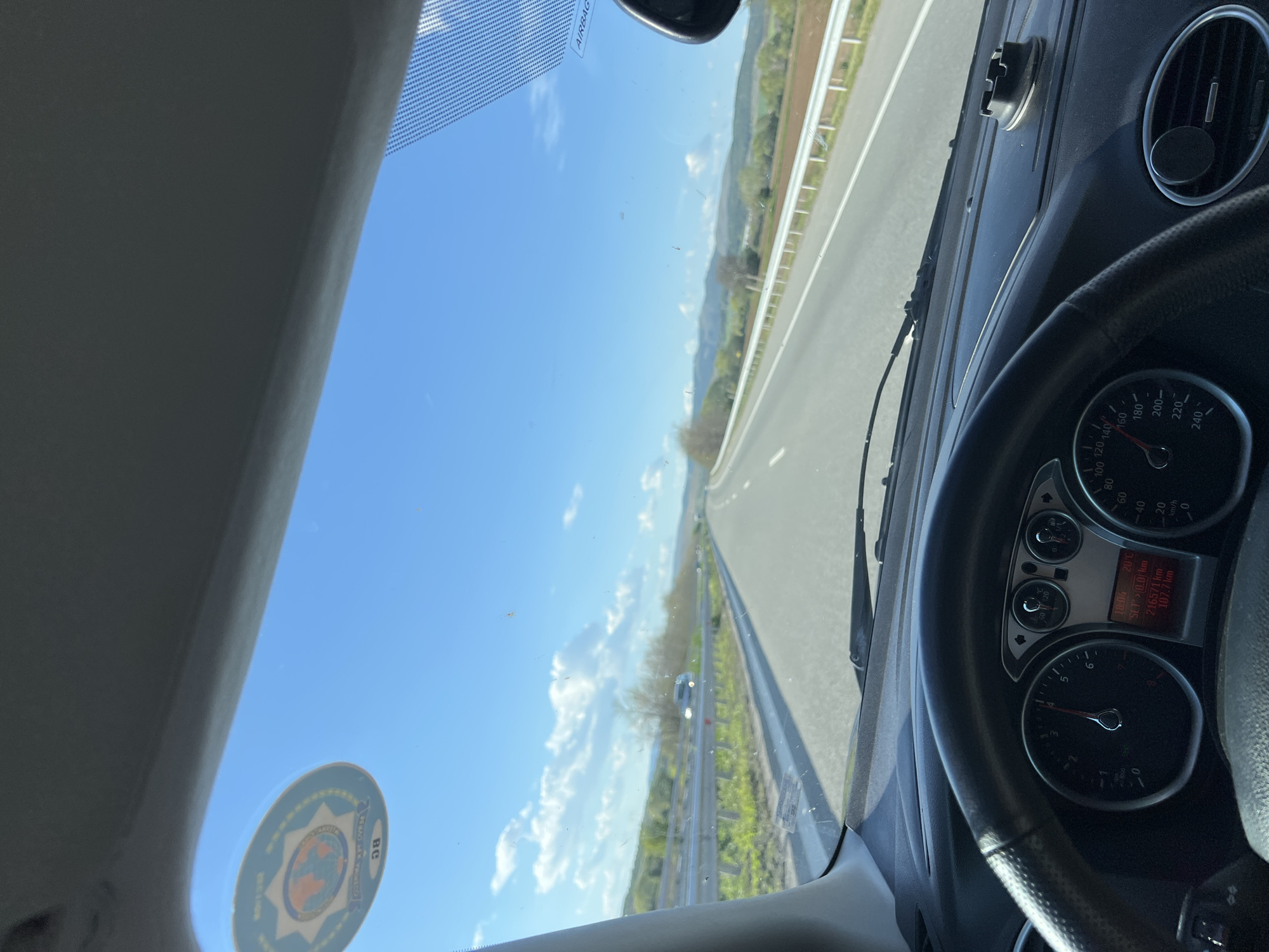 The image shows the view from inside a car driving on a highway, likely in Dolna Dikanya, Bulgaria, as suggested by the presence of a Bulgarian road tax sticker on the windshield. The speedometer reads approximately 5 km/h, and the odometer shows 216571 km. The exterior environment includes a blue sky with scattered clouds, a multi-lane highway with a center median barrier, and green vegetation along the roadside. There is another vehicle visible further down the highway.