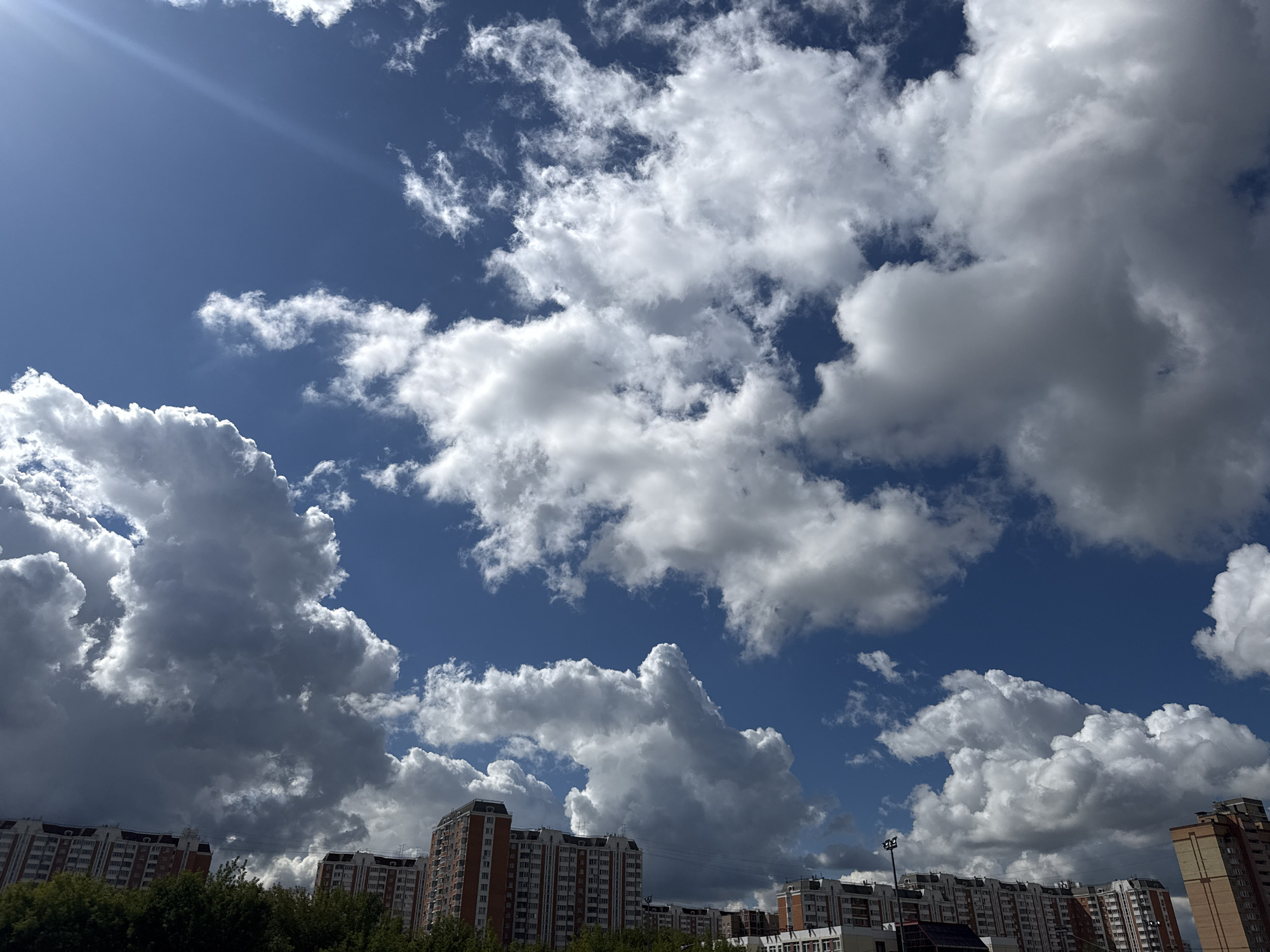 The image captures a view of the sky and buildings in Lyubertsy, Russia. A bright blue sky is filled with a variety of clouds, ranging from fluffy white cumulus clouds to darker, more ominous storm clouds. Sunlight beams down from the upper left, casting a lens flare across the sky. In the foreground, several multi-story residential buildings are visible, characterized by their orange and white facades. A line of green trees separates the buildings from the sky. The architecture suggests a post-Soviet urban environment. There are no people or animals visible in the scene. The overall impression is one of a dynamic sky over an urban landscape.