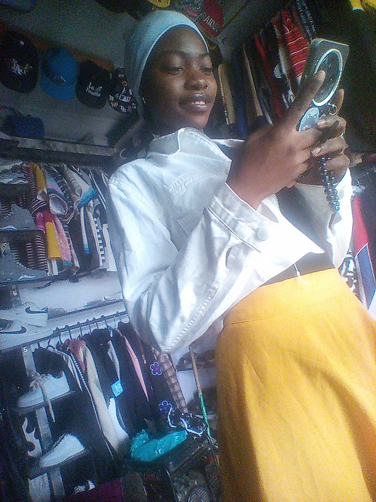 In an indoor setting, likely a clothing stall in Bukuru, Nigeria, a young person, wearing a light blue headscarf, a white jacket, and a yellow skirt, is holding a smartphone and a string of blue beads. The individual's right hand is raised, operating the phone, while their left hand holds the beads. The background is filled with clothing on racks, including hats and shirts. The lighting is dim, casting shadows.
