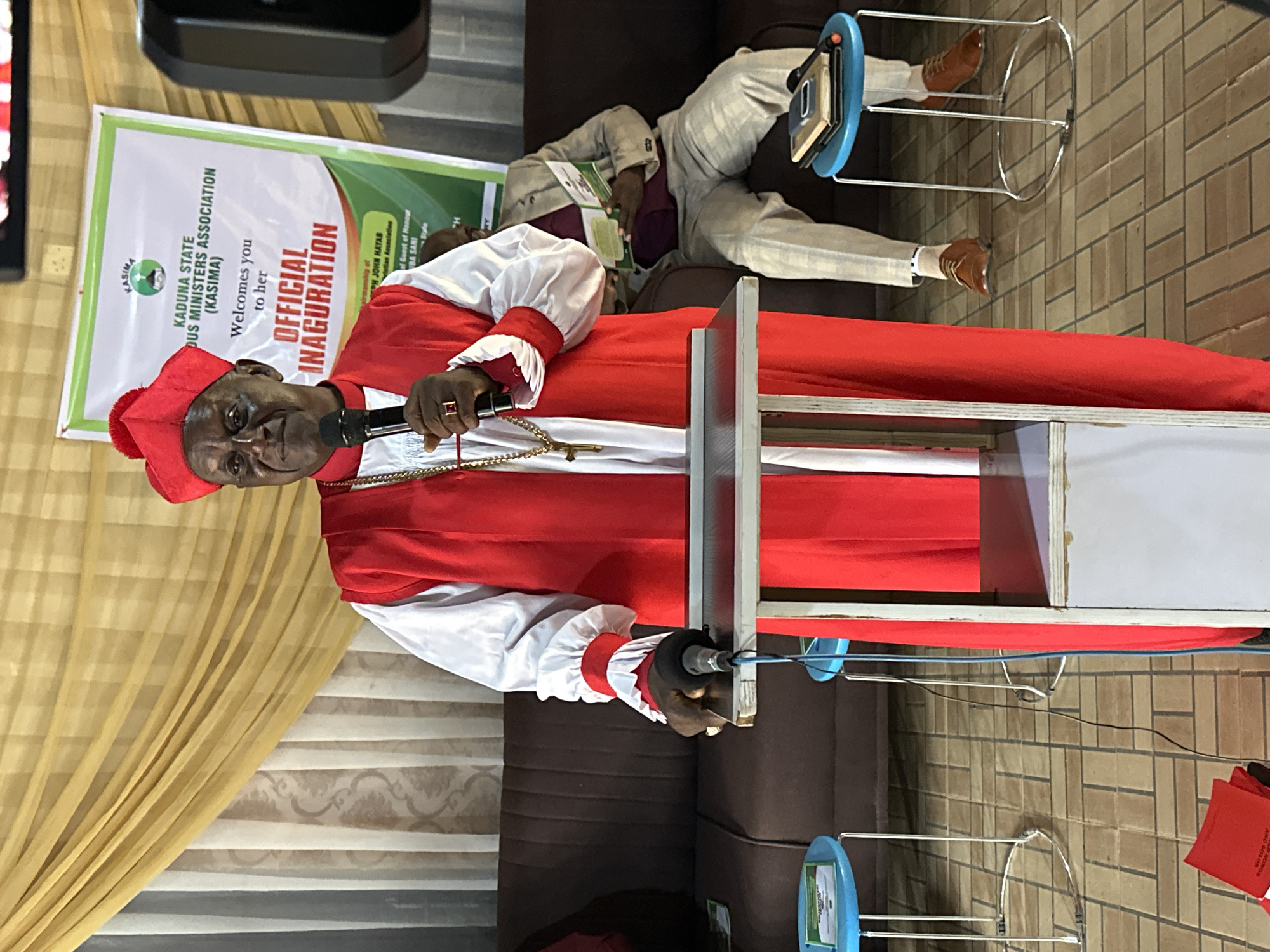A Black man wearing a red and white clerical robe and a red cap stands behind a podium, holding a microphone. He is at a podium with a microphone attached to it. Behind him, a banner reads "KADUNA STATE MINISTERS ASSOCIATION (KASIMA) OFFICIAL INAUGURATION". To the right of the speaker, a man in a light-colored suit sits on a couch, reading a booklet. The room has a tiled floor and is decorated with curtains. The scene appears to be from an official event in Barden Yana, Nigeria, possibly an inauguration or conference.