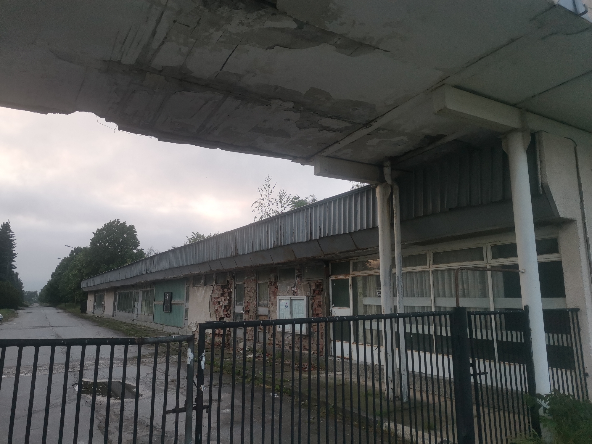 The image displays an abandoned or derelict industrial or administrative building located in Zhivkovo, Bulgaria. The long, low-rise concrete structure exhibits extensive signs of decay, particularly noticeable on the overhanging flat roof section in the foreground, which shows severe cracking, spalling, and missing concrete. Several white concrete columns support this deteriorating canopy.
The building's facade is in disrepair, with areas of exposed brickwork where plaster or render has fallen away, and peeling paint. Multiple rectangular windows, some partially obscured by white curtains, line the building, while others appear boarded or broken. A corrugated metal roof covers the main building.
In the immediate foreground, a black metal gate with vertical bars partially blocks the view, suggesting a secured or enclosed property. Beyond the gate, an asphalt road stretches into the distance, featuring puddles that indicate recent rainfall. The road is lined on both sides by a mix of coniferous and deciduous trees, enhancing the sense of desolation.
The sky is overcast, lending a dull, diffused light to the scene, possibly indicating early morning, late afternoon, or simply a very cloudy day. No people are visible, and no activity is taking place, contributing to the overall impression of abandonment and neglect. Through a window on the right side of the building, a faint sign with indecipherable text can be glimpsed, a minor detail in the larger picture of decay.