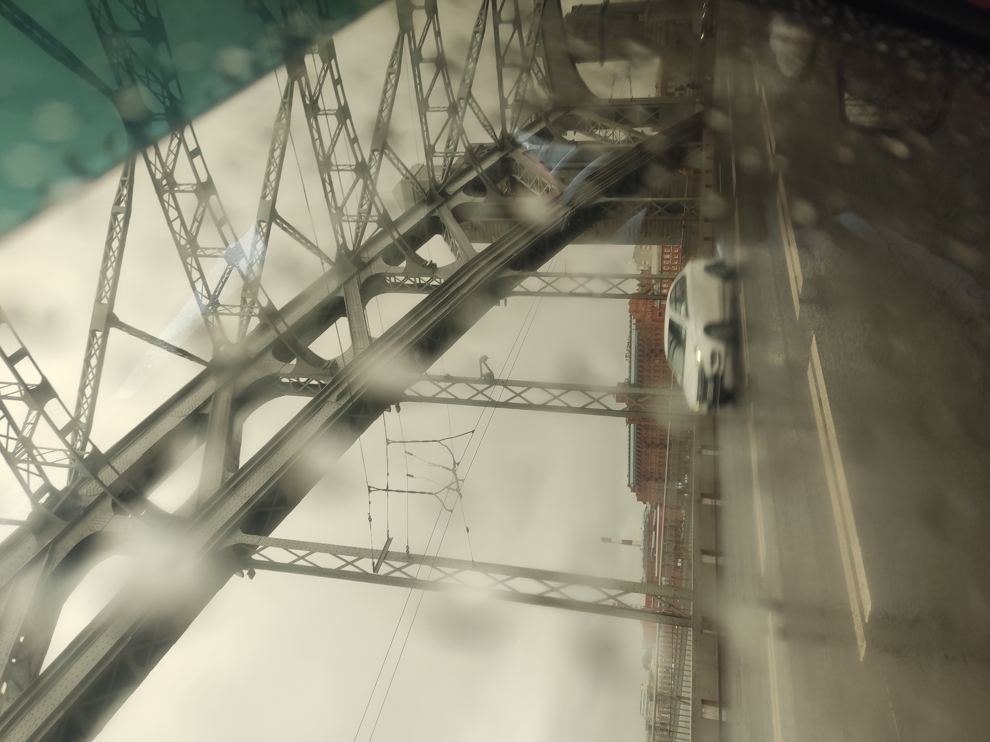 A view through a vehicle windshield, heavily obscured by raindrops, shows the Peter the Great bridge in Saint Petersburg, Russia. The bridge's steel structure dominates the left side of the frame. A white car is visible on the roadway, traveling on the right. The sky is overcast, creating a grey and muted light. A few brick buildings are also seen, with smokestacks.