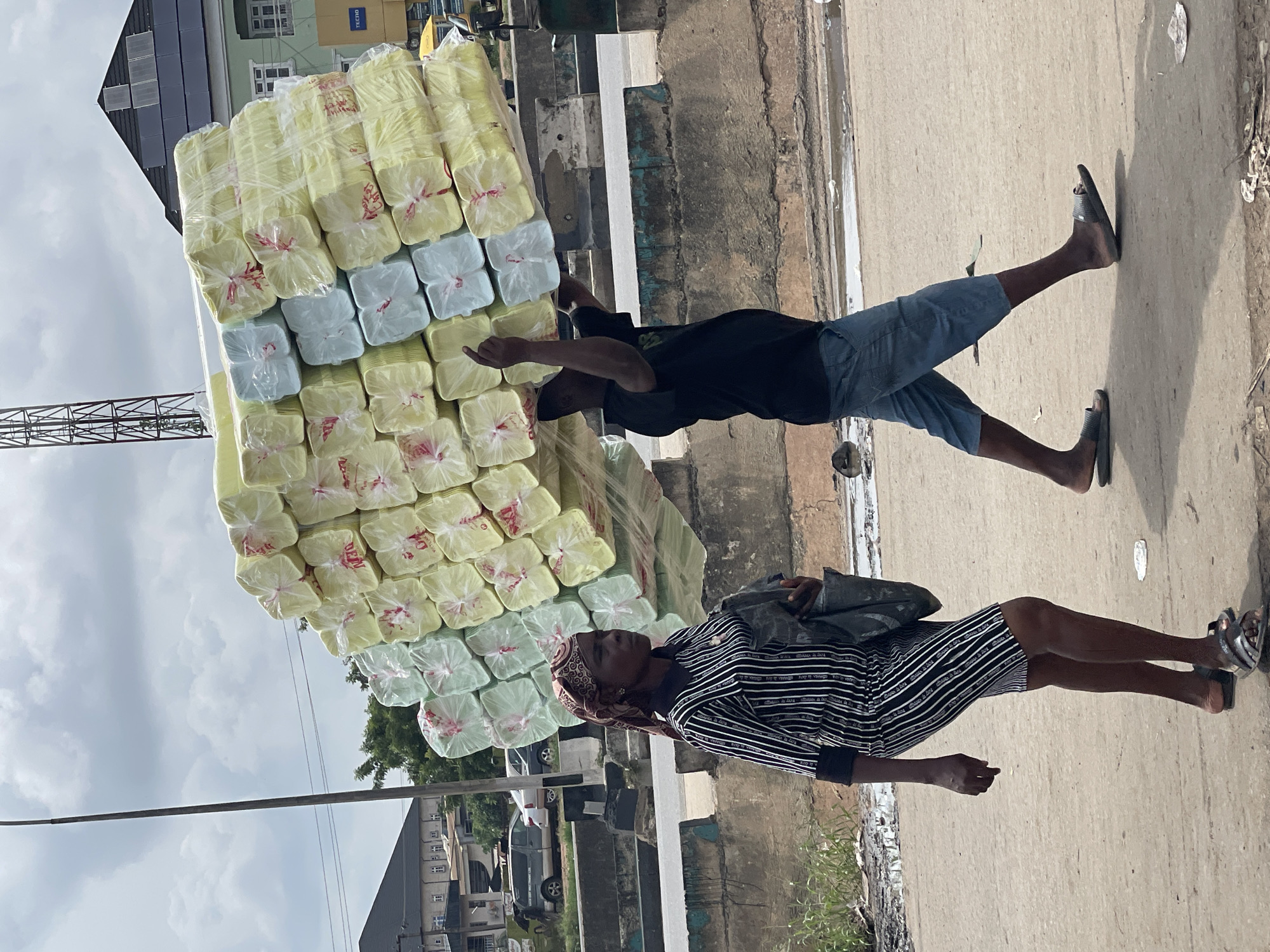 A man and a woman walk on a paved surface in an urban environment, carrying a large, stacked load of light yellow and light blue foam sheets wrapped in clear plastic. The man, in the foreground and to the right, is carrying the majority of the load on his head, balanced with his arms. He is wearing a black t-shirt and blue shorts. The woman is walking to the left of the man and slightly behind him, carrying a dark bag over her shoulder. She is wearing a black and white striped dress and a headscarf. In the background, there are buildings, a street, and a cloudy sky. The overall scene appears to be a busy street in a city, possibly a market area given the large quantity of goods being transported.