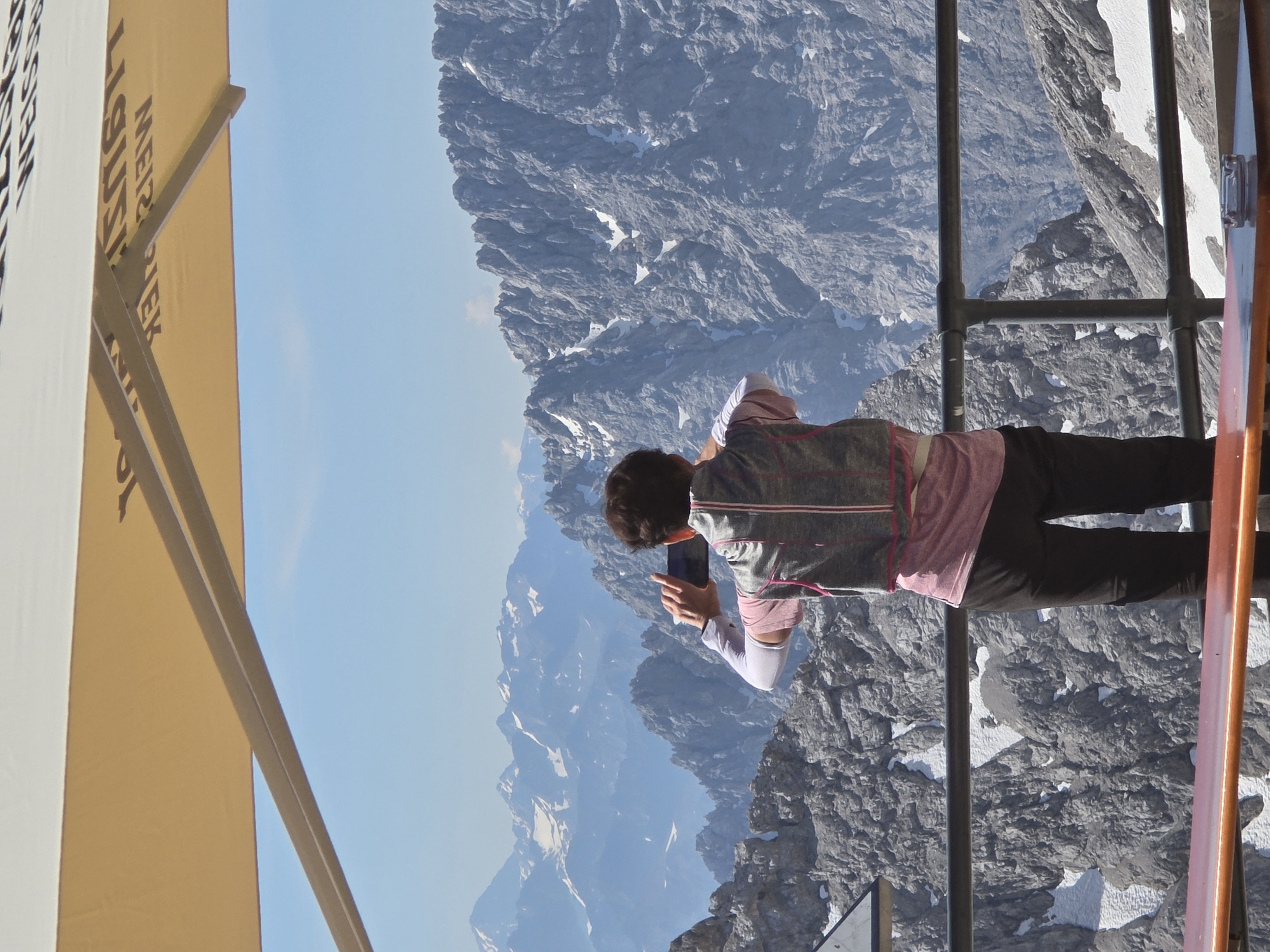 A person stands on a balcony overlooking a vast, rugged mountain range with snow-capped peaks. The person is facing away from the camera, holding a smartphone up to take a photograph of the landscape. They are wearing a grey vest over a pink shirt and black pants, with white arm sleeves. A wooden table with an ashtray sits in the foreground. A large tan umbrella provides shade for the balcony. The scene appears to be at a high altitude, possibly at the Zugspitze, Germany's highest peak, which is near Garmisch-Partenkirchen. The sky is clear and blue, indicating good weather. The mountains are predominantly rocky and grey, with patches of white snow visible on their slopes. The overall impression is one of a breathtaking natural vista and a moment of personal engagement with it.