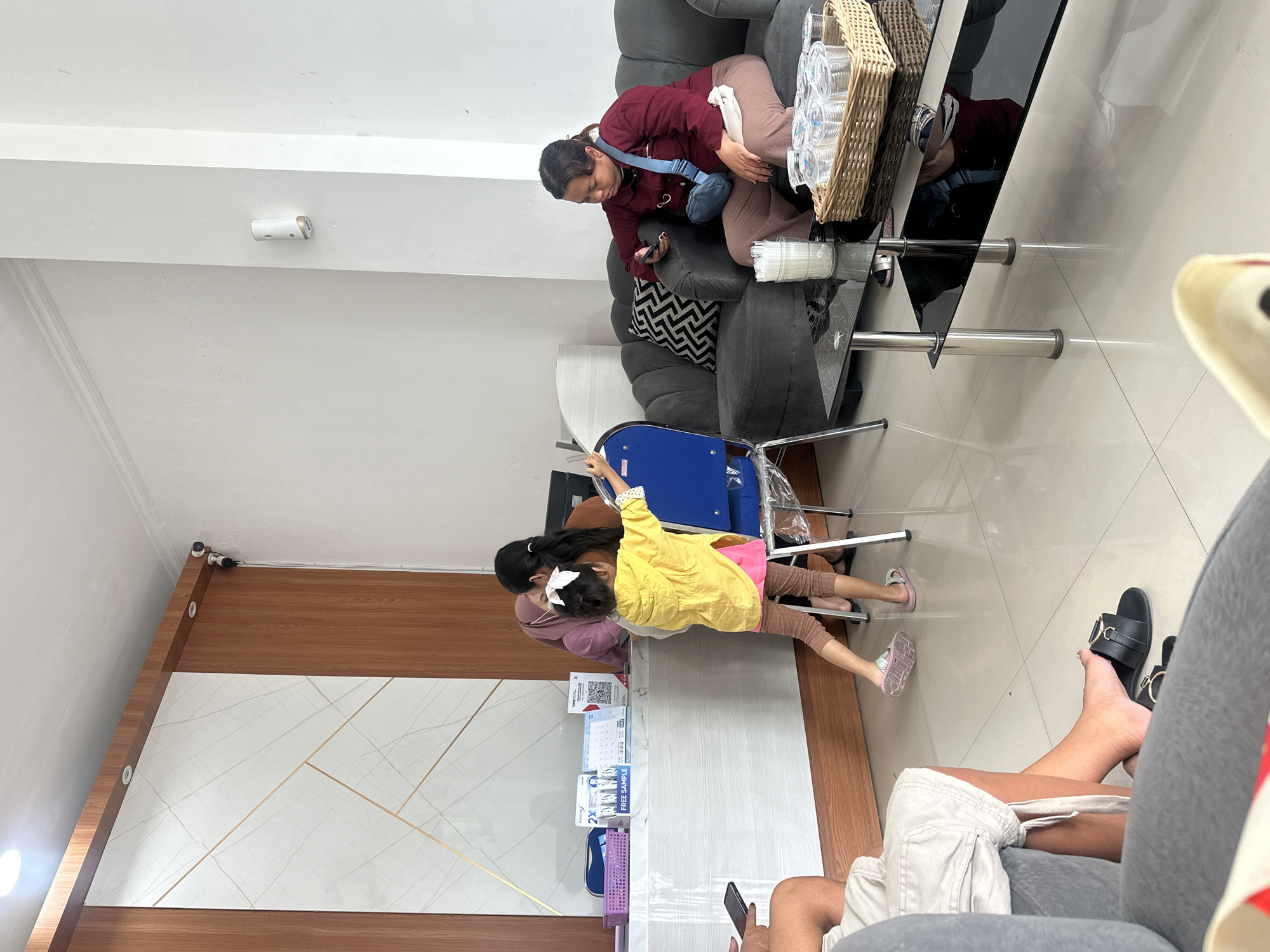 The scene depicts a waiting area in what appears to be a business in Denpasar, Indonesia. A woman is seated on a grey couch, looking at her mobile phone, while a basket of plastic cups sits on a glass-topped table nearby. A girl in a yellow jacket interacts with someone at a counter that features a display of promotional materials and a calendar. The wall behind the counter is decorated with geometric patterns. Other individuals are present, one appears to be using a mobile phone, and footwear is visible. The environment is indoors and relatively bright.