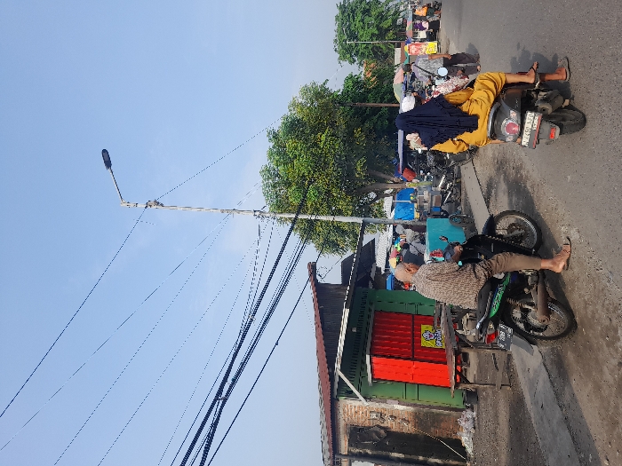 Two motorcycles are parked on the side of a road. One has two people on it, the passenger wearing a head covering. The other motorcycle has a single rider, also wearing sandals. A building is in the foreground with a red and green facade. Overhead power lines cross the sky towards a street light. Trees are in the background, and some tents are pitched nearby.