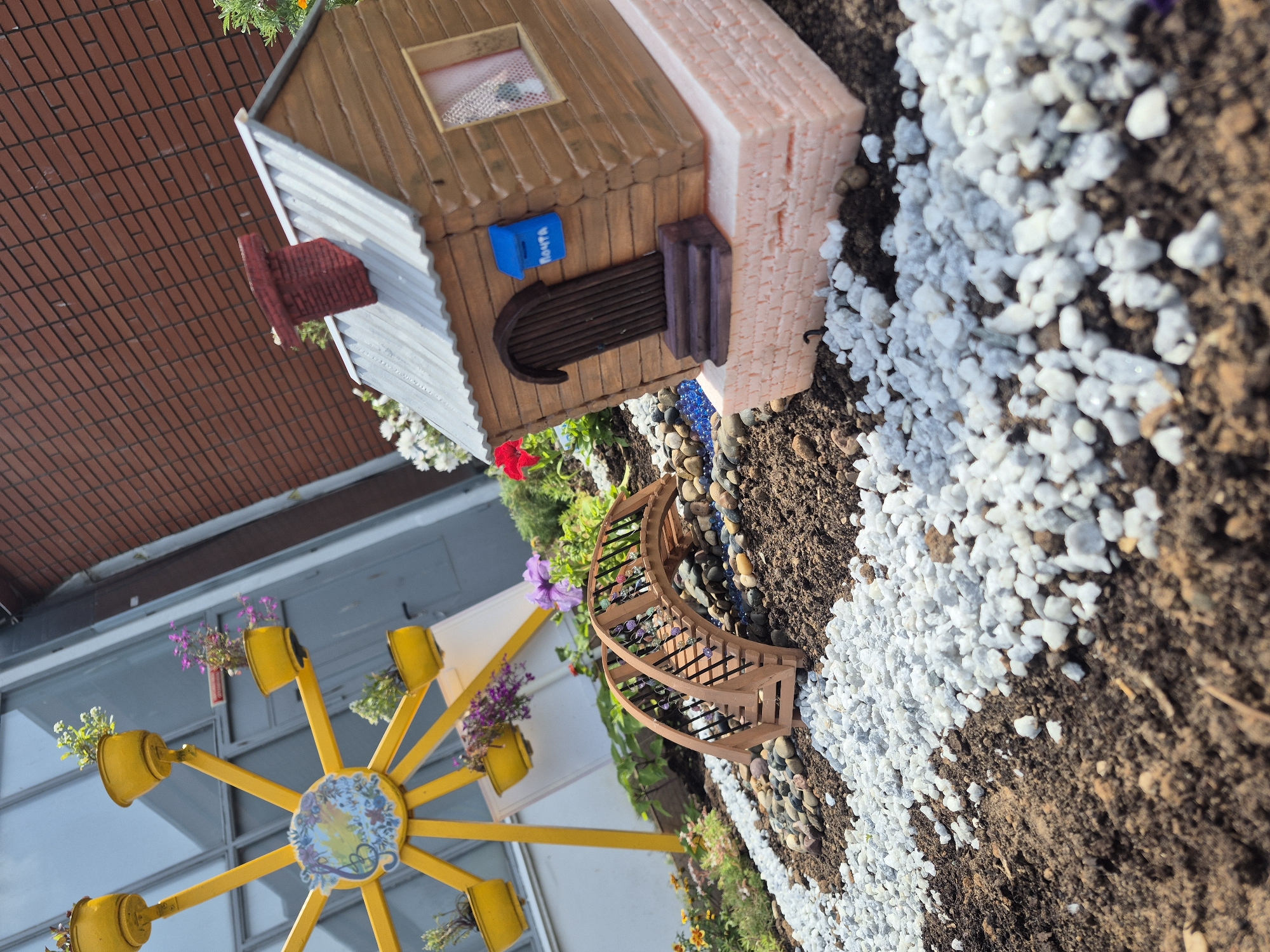 This media file depicts an outdoor scene with miniature elements, possibly part of a garden or display. The main subjects include a small, toy-like house with a brown wooden exterior and a silver corrugated roof. The house features a small window with lace curtains, a dark brown arched doorway, and a blue mailbox with the word "ПОЧТА" (Posta, meaning "Mail" in Russian) visible. Adjacent to the house are decorative elements like a small wooden bridge with railings and a yellow Ferris wheel-shaped structure adorned with potted plants and flowers. The ground is covered with dark soil and decorative white pebbles. In the background, a brick wall is visible, suggesting an urban or semi-urban setting. The lighting suggests it is daytime, with shadows indicating sunlight. The overall scene evokes a whimsical or playful atmosphere, possibly representing a miniature village or a creative garden design.