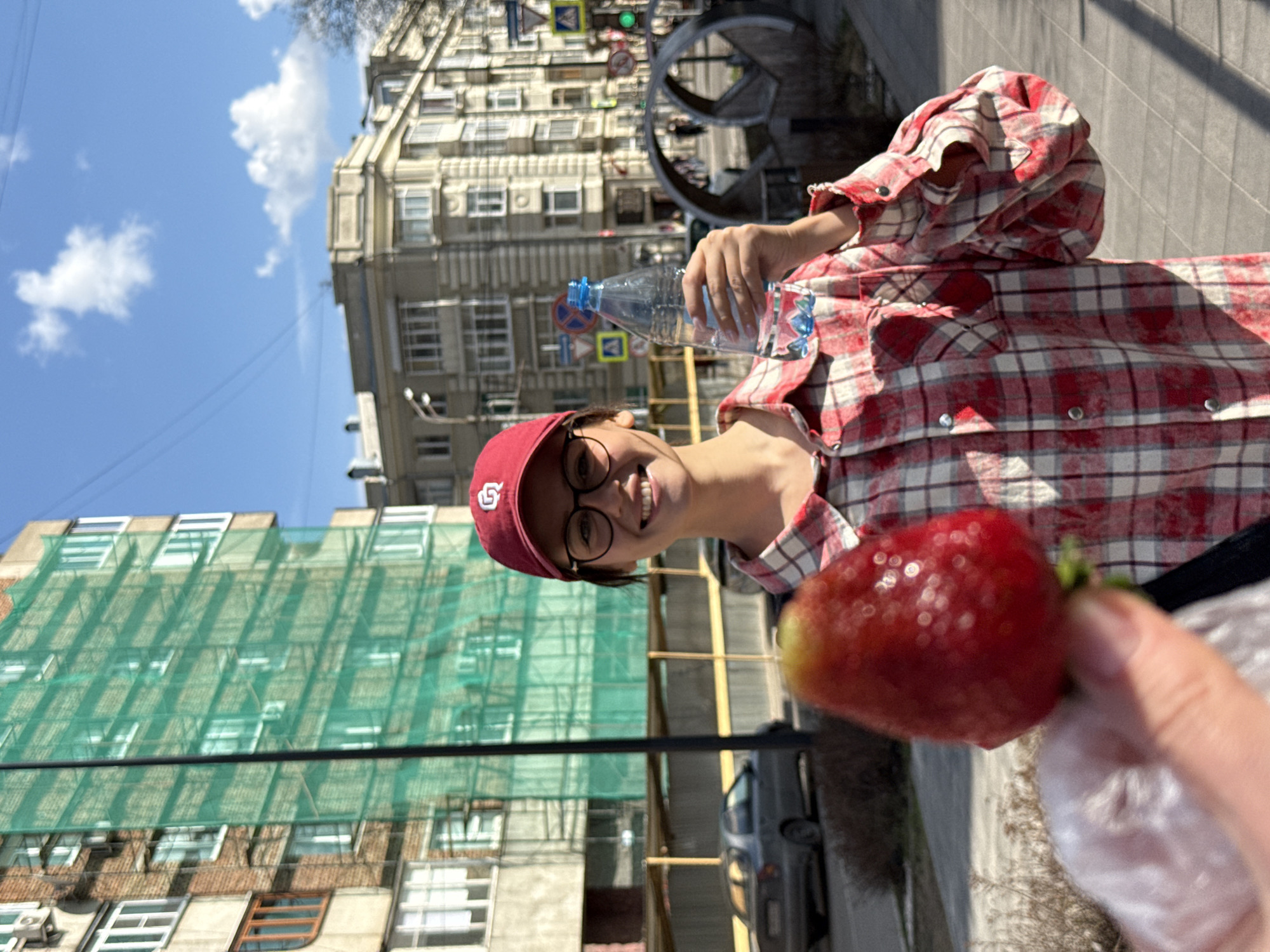A street-level medium shot depicts a person holding a large strawberry in the foreground. The person is light-skinned, wearing black-rimmed glasses and a red baseball cap, smiling. They are wearing a red and white plaid shirt and holding a plastic water bottle.
The background shows a cityscape. To the left is a building with a green mesh covering its facade, likely for construction or renovation. Further back, another building with light-colored facade is visible. A street is visible in the lower-middle ground with cars parked along the curb. Overhead traffic signals and a metal structure, possibly a decorative arch, are present in the background. The sky is blue with light clouds.
Based on visual clues and known geographical locations, the photo was taken in Krasnoyarsk, Russia. The buildings and streetscape are consistent with the urban environment of this city.