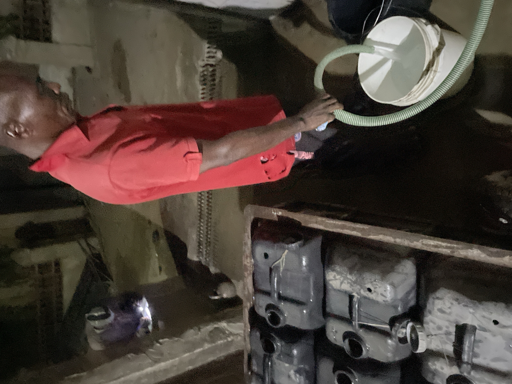 An individual wearing a red shirt is holding a green hose connected to a white bucket, appearing to pour liquid into a set of dark containers. These containers are stacked and housed within what seems to be a concrete or metal frame. The scene is dimly lit, suggesting an indoor or nighttime setting in an urban environment. The location is listed as Unknown, Unknown.