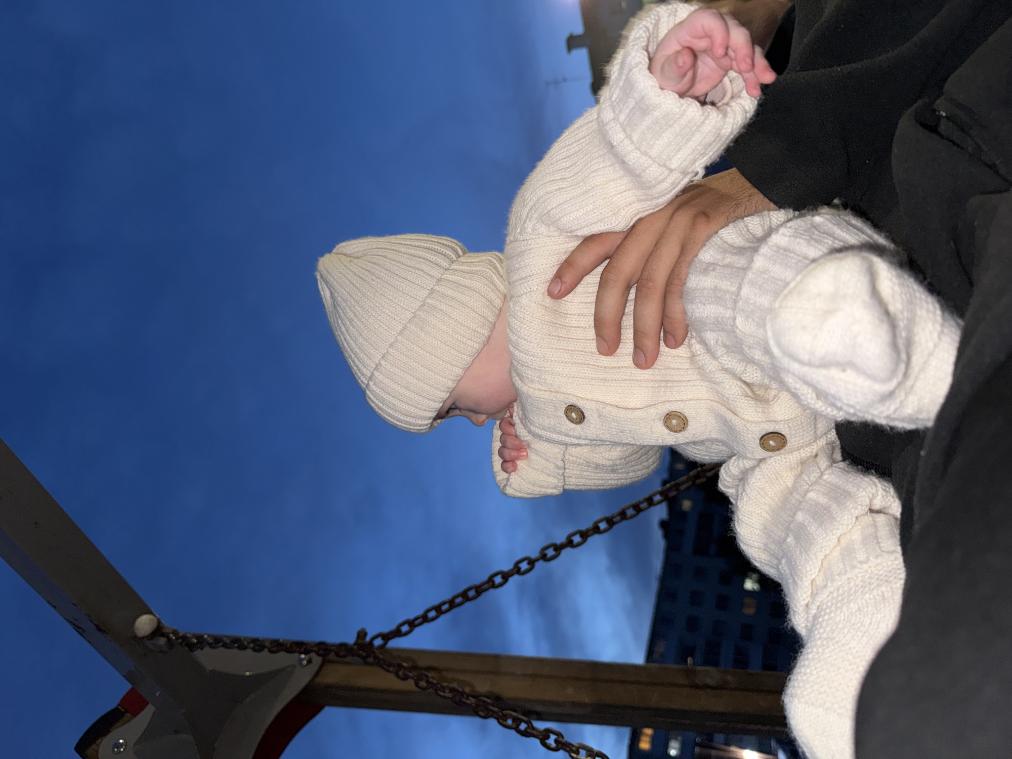 An outdoor, low-angle, medium shot captures a baby in a cream-colored knitted onesie and hat, held by an adult. The baby faces right, with their head tilted down, and appears to be sucking on their hand. The adult's hands cradle the baby. Behind them, a chain is attached to a swing set structure on the left, and in the background, a dark blue twilight sky hovers above a dimly lit apartment building. The scene is set in Noviy, Russia.