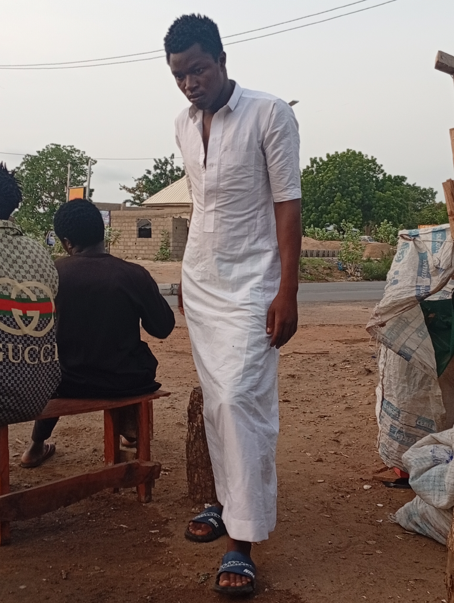 The image shows a man standing in an outdoor setting in Adankolo, Nigeria. He is wearing a long, white, robe-like garment and blue sandals. To the left of the man, two individuals are sitting on a wooden bench. The person closest to the camera wears a patterned shirt with the word "GUCCI" visible. Next to the man, there are white bags or sacks. The background shows a dirt or gravel ground, some trees, and a building. A road is also visible further back. The overall environment appears to be somewhat rural or a roadside area.