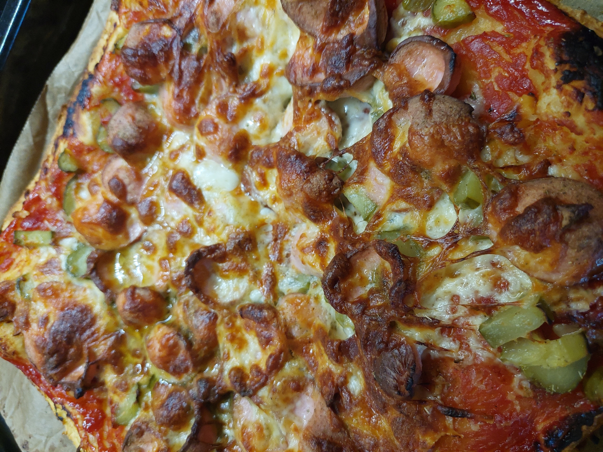 A close-up shot shows a square pizza, likely homemade, resting on baking paper. The toppings are unevenly distributed, with one half appearing to have a white sauce base and the other a red tomato sauce base. Both sides are topped with sliced sausages and diced pickles. The cheese is melted and bubbly, and the crust is slightly burnt in places. The location is unknown.