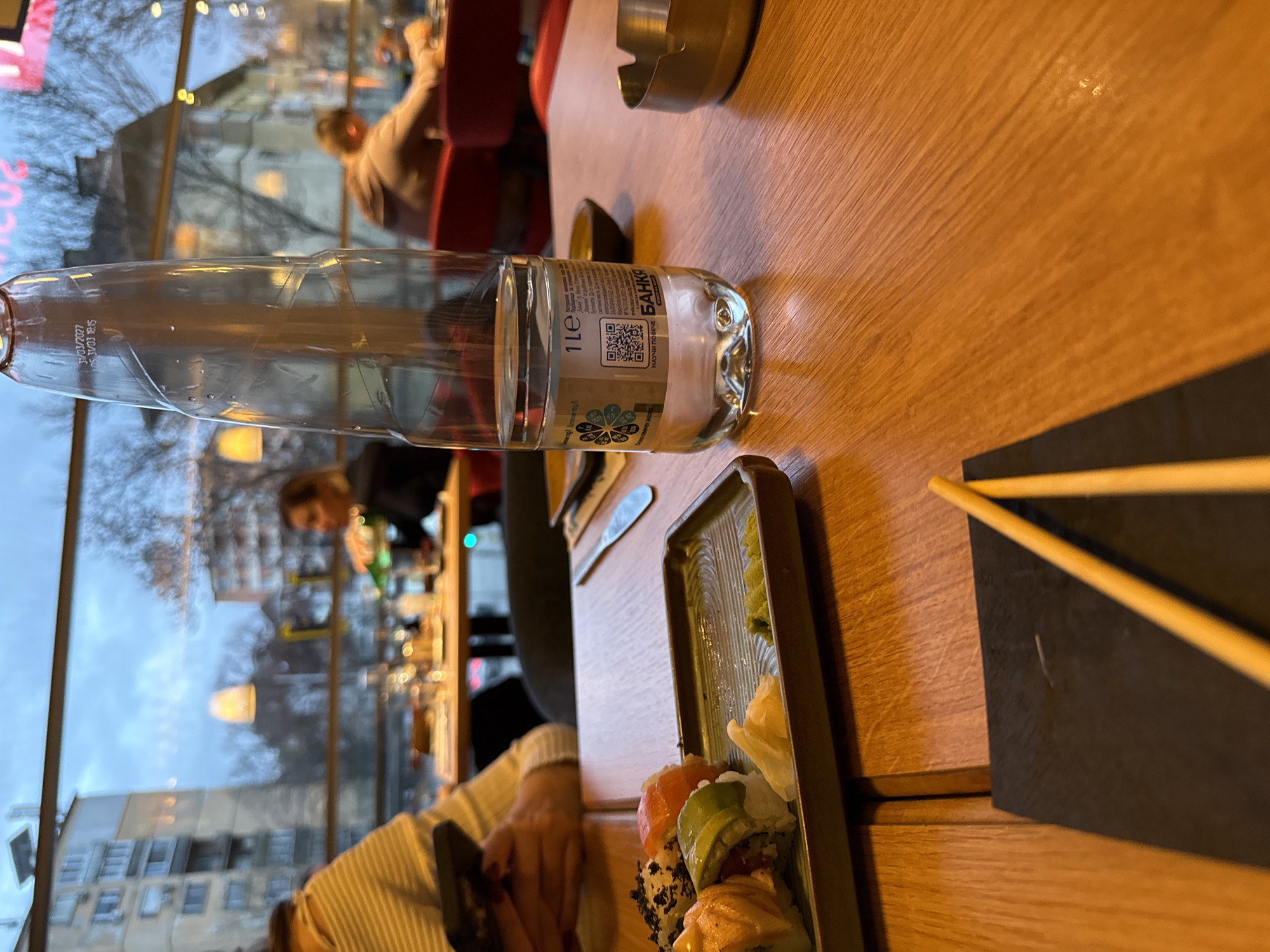 A restaurant scene in Sofia, Bulgaria. A wooden table holds a bottle of Банкя mineral water, an ashtray, and sushi on a dark green tray. Chopsticks rest on a black placemat. In the background, a large window reveals a cityscape with buildings and leafless trees. Patrons are visible in the restaurant. One is partially seen in the foreground, holding a dark object, and others are at tables near the window. The light suggests it may be late afternoon or evening.