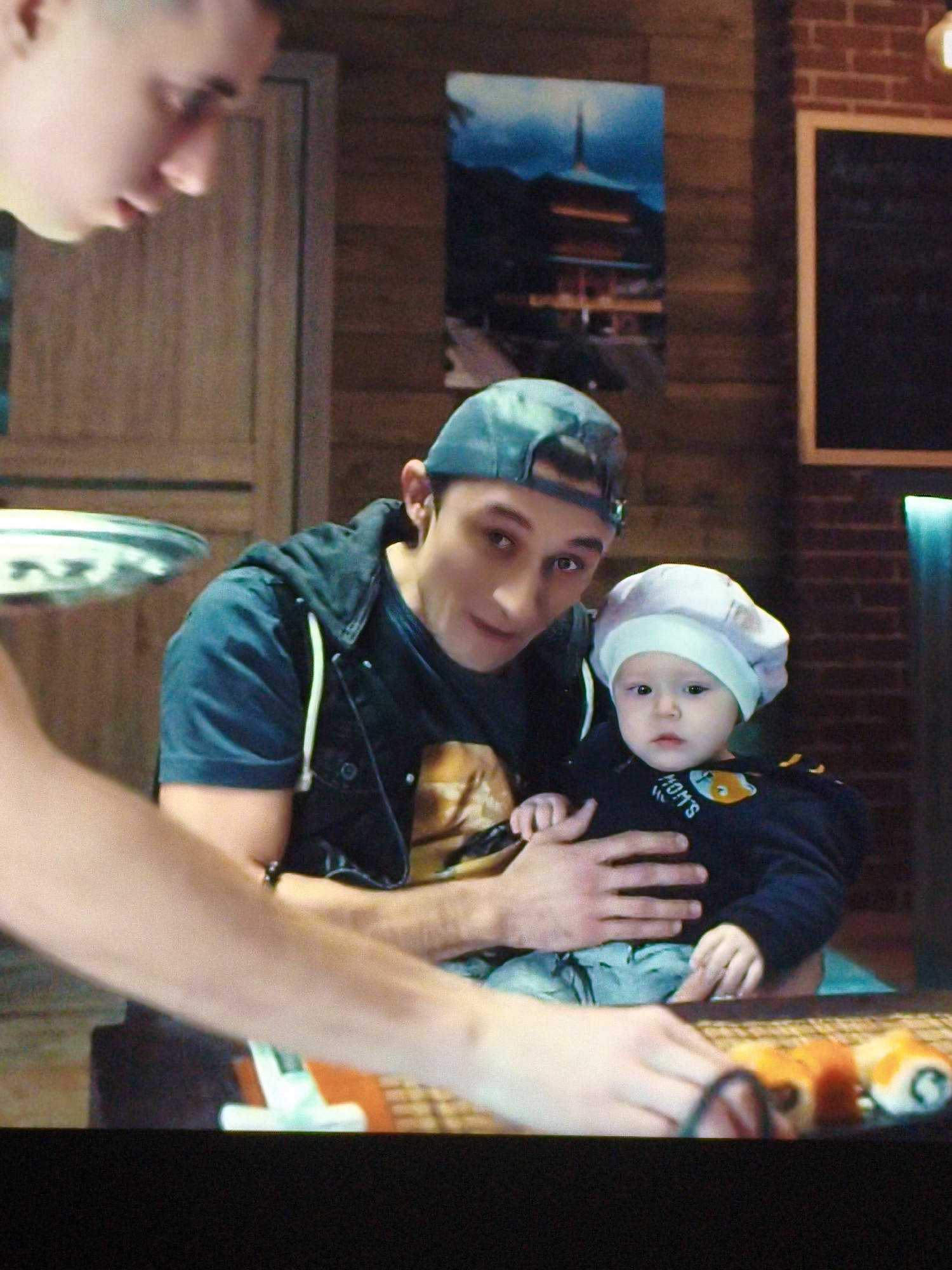 An adult male wearing a blue baseball cap and a dark hooded vest over a t-shirt is holding a baby. The baby is wearing a light-colored bonnet and a dark long-sleeved shirt with "MOM'S" written on it in white letters. They are seated at a table with sushi rolls and billiards balls visible. The background features wooden paneled walls, a framed picture of a pagoda, and a chalkboard. The scene does not provide any specific clues to indicate the location is Vladivostok, Russia.