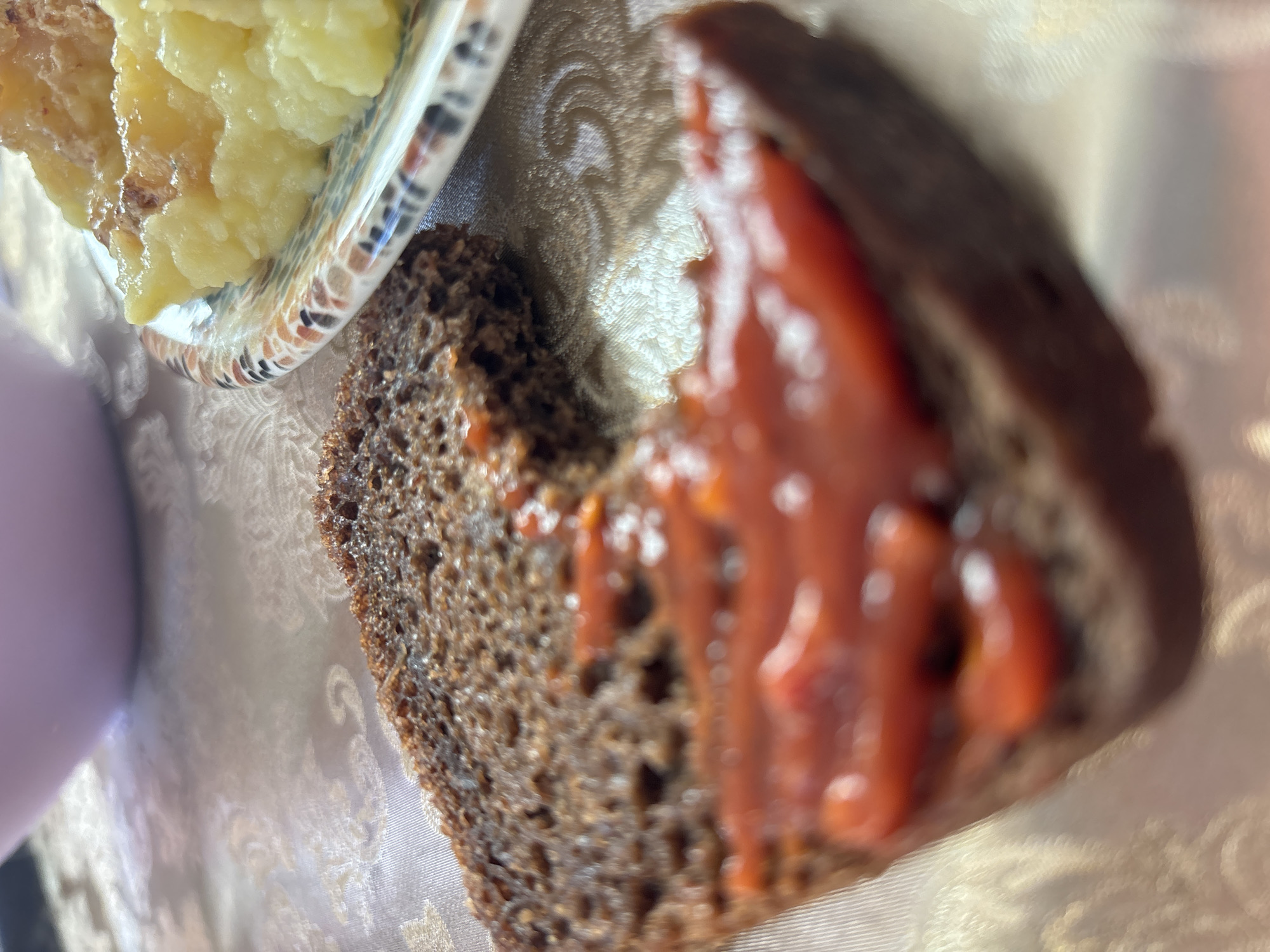 A close-up, shallow-depth-of-field shot shows a slice of dark bread topped with a red sauce, likely tomato paste or ketchup. The bread is slightly charred at the edges. To the right, a plate with a colorful, patterned rim holds a portion of a yellow, mashed food, possibly potatoes or a corn puree. In the background, a blurred purple bowl or dish is visible. The food is set on a light-colored tablecloth with a subtle, ornate pattern. The scene suggests a simple meal in a domestic setting within Vsevolozhsk, Russia.