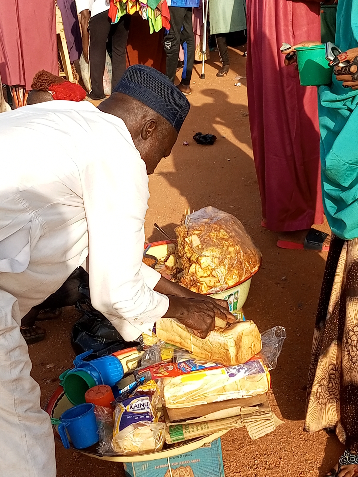 The image shows a market scene in an outdoor setting. A man wearing a white robe and a dark blue cap is bent over a table, interacting with the items on it. His hands are visible, and he appears to be handling food items and some packaged goods. On the table, there are loaves of bread, plastic cups in various colors (blue, green, orange), and a large transparent bag filled with a golden-brown, crunchy food item, possibly snacks or fried dough. Some packaged goods, including what appears to be a loaf of bread wrapped in plastic with the label "KAINU" and other food items, are also present. Behind the man, other people are visible in the background, some wearing colorful clothing. The ground is sandy and appears to be dirt. The overall impression is of a busy marketplace.