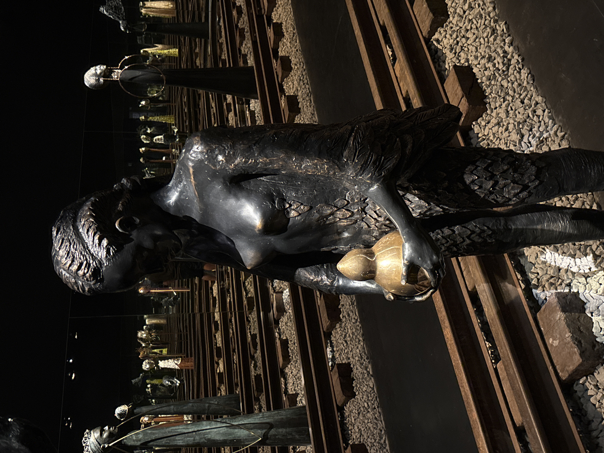 The image depicts a bronze statue of a woman with feathered skin, holding a golden orb. The statue is positioned along a railroad track lined with gravel and exposed wooden ties. The environment suggests an art installation within a dark or dimly lit space, potentially a museum or gallery. The reflections of other statues and objects are visible in the background, giving an impression of a mirrored exhibit. The location is in the city of São Paulo, Brazil, based on the general style of art installations and sculptures commonly found in its public spaces and museums.