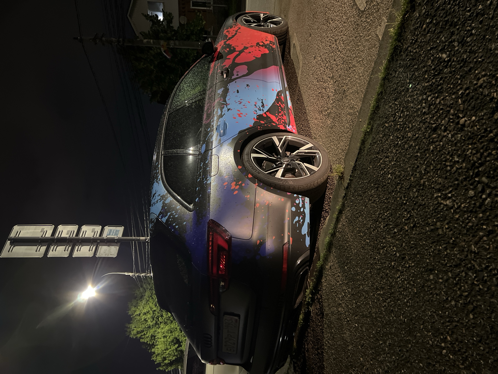 A dark-colored Audi A4 Avant wagon is parked on an asphalt road at night, in what appears to be a residential area in Krasnodar, Russia. The car has a custom wrap with a colorful, abstract design featuring splashes of red, blue, and pink against a dark background. The vehicle's rear is visible, showing a license plate that reads "E029PT 193". A street light illuminates the scene, casting a bright glow and creating lens flare. Trees and a building are visible in the background. The road is wet, suggesting recent rain.