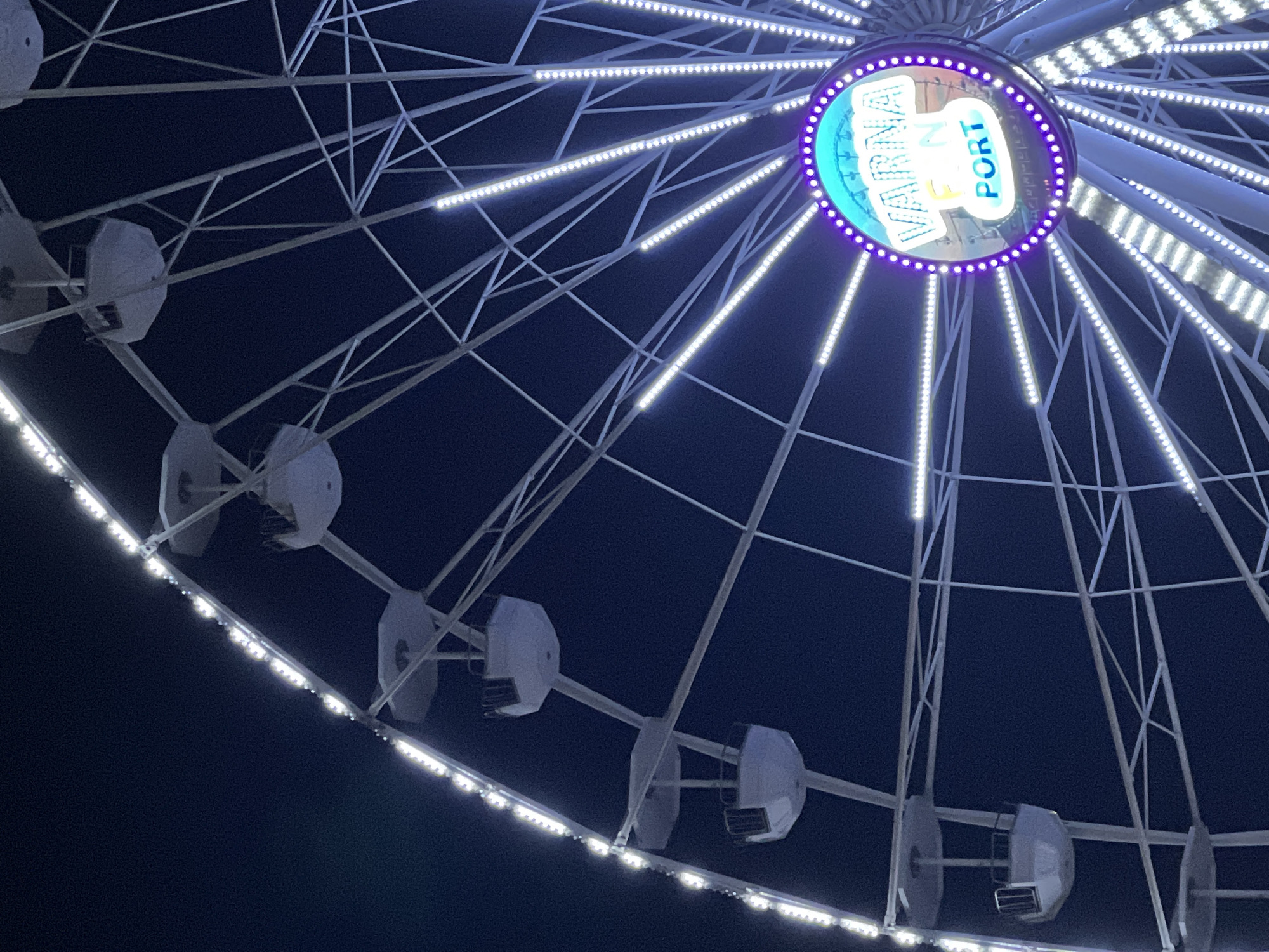 The image displays a brightly illuminated Ferris wheel against a dark, clear night sky, strongly indicating it is nighttime. The large structure features an intricate network of white metal spokes radiating from a central hub. Numerous white LED strip lights adorn these spokes and portions of the wheel's outer rim, creating a striking visual effect.
The central hub of the Ferris wheel is prominently lit with a distinctive logo. The dominant text visible on this circular hub reads "VARNA" in large, light-colored letters, set against a background that appears to be a light blue or teal. Below and to the right of "VARNA," the word "PORT" is also clearly legible. The entire central logo is outlined by a ring of vivid purple lights, providing a colorful contrast to the white lights elsewhere on the wheel.
Several enclosed, light-colored cabins are visible hanging from the Ferris wheel's structure, suggesting it is a passenger attraction. These cabins appear empty in the frame. The scene conveys the impression of a vibrant amusement or entertainment attraction, illuminated for display or operation during the evening. Given the text "VARNA PORT," the location is Varna, Bulgaria, likely situated near its port.