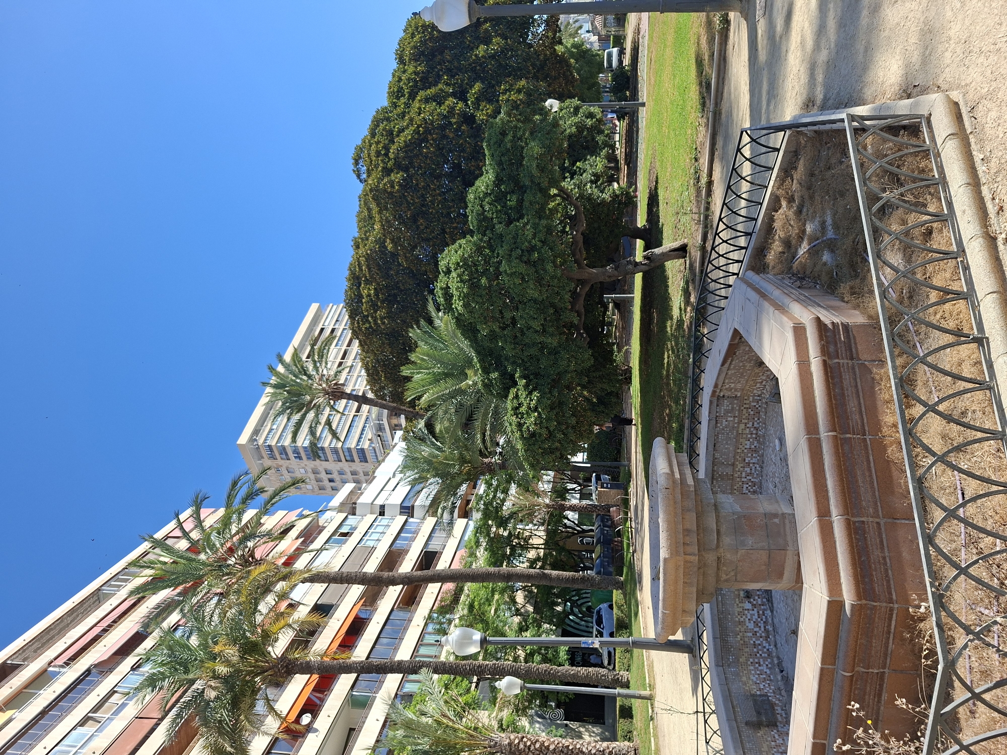 The image depicts a sunny day in a park in Alicante, Spain. In the foreground, a dry, ornate stone fountain with mosaic tile accents is enclosed by a decorative metal fence. Beyond the fountain, a paved pathway leads into a grassy area with several palm trees and a large, dense tree. In the background, tall apartment buildings with balconies and awnings rise against a clear blue sky. A few cars and streetlights are visible in the mid-ground, and the ground is covered with dirt paths and green grass. The scene is peaceful and appears to be a public urban space.