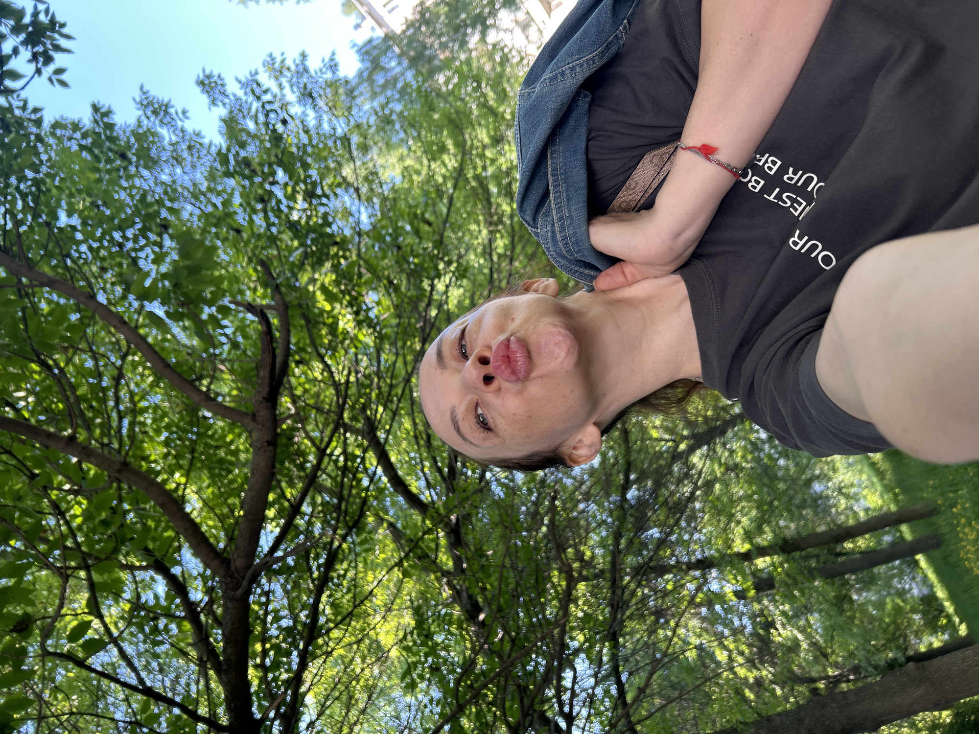 The image shows a selfie taken from a low angle, looking upwards at a woman making a kissing face. Behind her are tree branches and green leaves, with glimpses of a blue sky. A building is partially visible in the upper right corner. The woman is wearing a dark gray shirt with white text, and a denim jacket is draped over her shoulder. A red bracelet is visible on her wrist. The location may be a park in the city of Chaikovsky, Russia.
