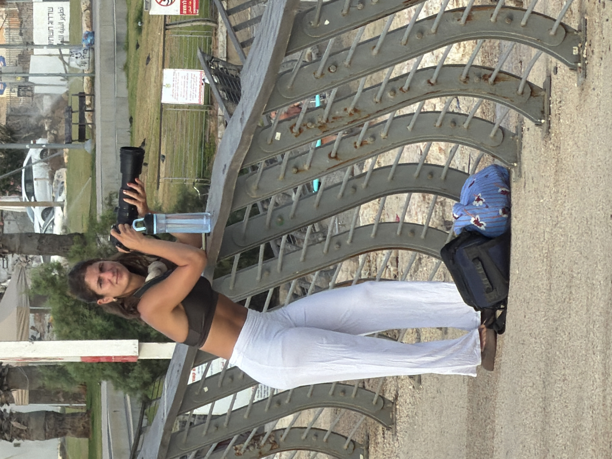 A woman, dressed in a dark sports bra and white flowing pants, stands on a promenade in Tel Aviv, Israel. She is holding a large camera with a telephoto lens and a blue water bottle rests beside her. She has headphones around her neck. In the background, a white car is parked on a street, and palm trees and vegetation are visible. The promenade has a metal railing and a paved surface. There are also some benches and signs in the distance. The woman is looking at the camera, as if posing.
