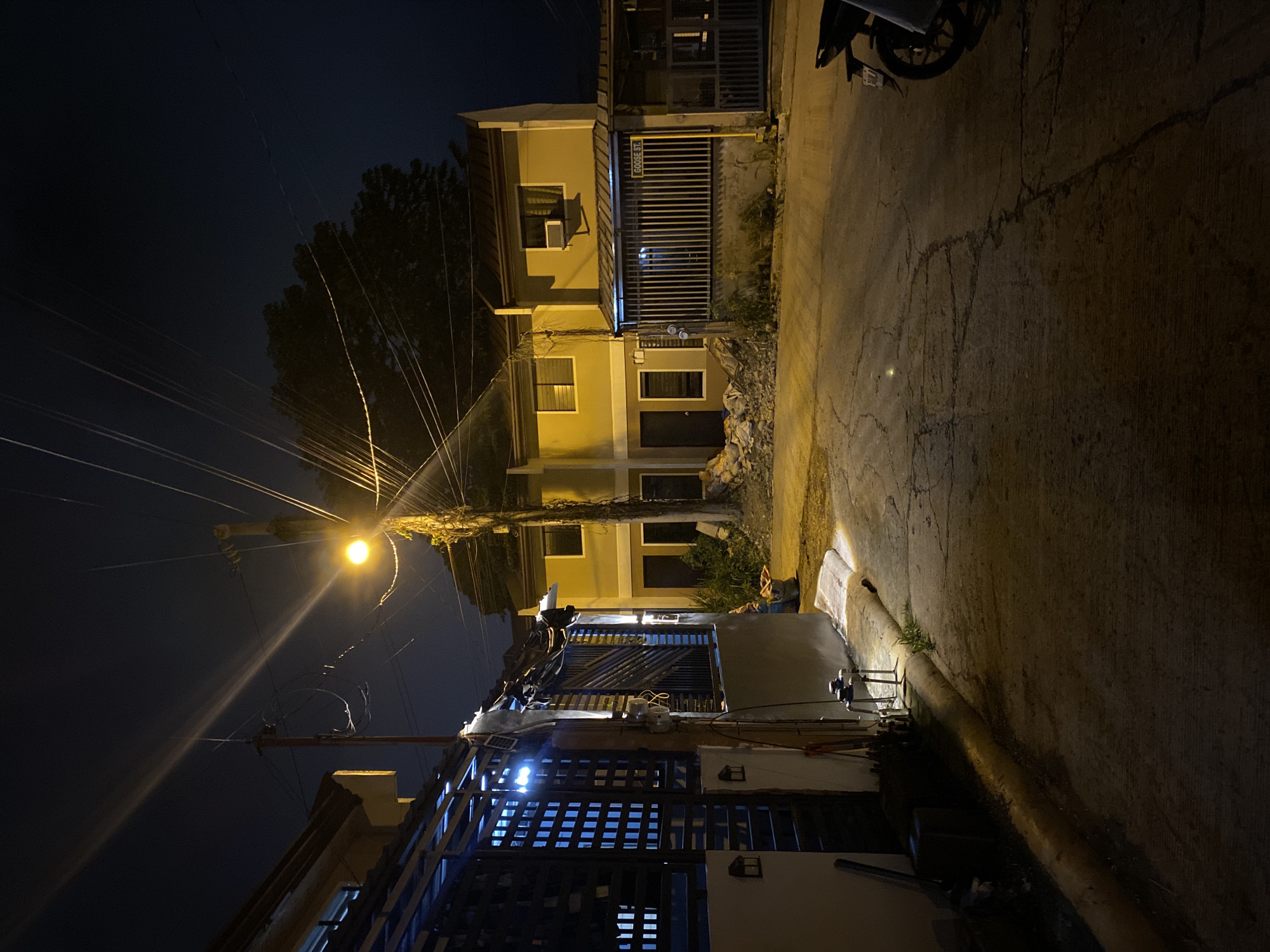 This media file captures a nocturnal street scene in what appears to be a residential area in Davao City, Philippines. The image is oriented vertically (landscape viewed as portrait) but depicts a narrow, concrete road flanked by two-story houses.
The primary light source is a bright, yellow street lamp mounted on a tall utility pole situated on the left side of the road. This pole is heavily overgrown with vines and supports a complex web of electrical and communication wires that crisscross the dark night sky.
On the right side of the road, two houses are visible. The upper house is light-colored (beige or light brown) with barred windows and an air conditioning unit. A prominent yellow sign with black text reading "GOOSE ST" is affixed to its gate. Below it, another two-story residence features a dark entrance and metal gates with visible blue-white interior or security lights. The ground in front of the houses shows piles of what looks like debris or trash.
On the left side of the road, glimpses of another property with patterned metal gates and white lights are visible. The road itself is made of concrete, showing numerous cracks and some patches of brighter light, possibly from vehicle headlights or other distant sources. A motorcycle or scooter is partially visible at the upper right edge of the frame (when the image is viewed in its original orientation). No people are visible, and no specific activity is taking place, presenting a quiet, illuminated street at night.