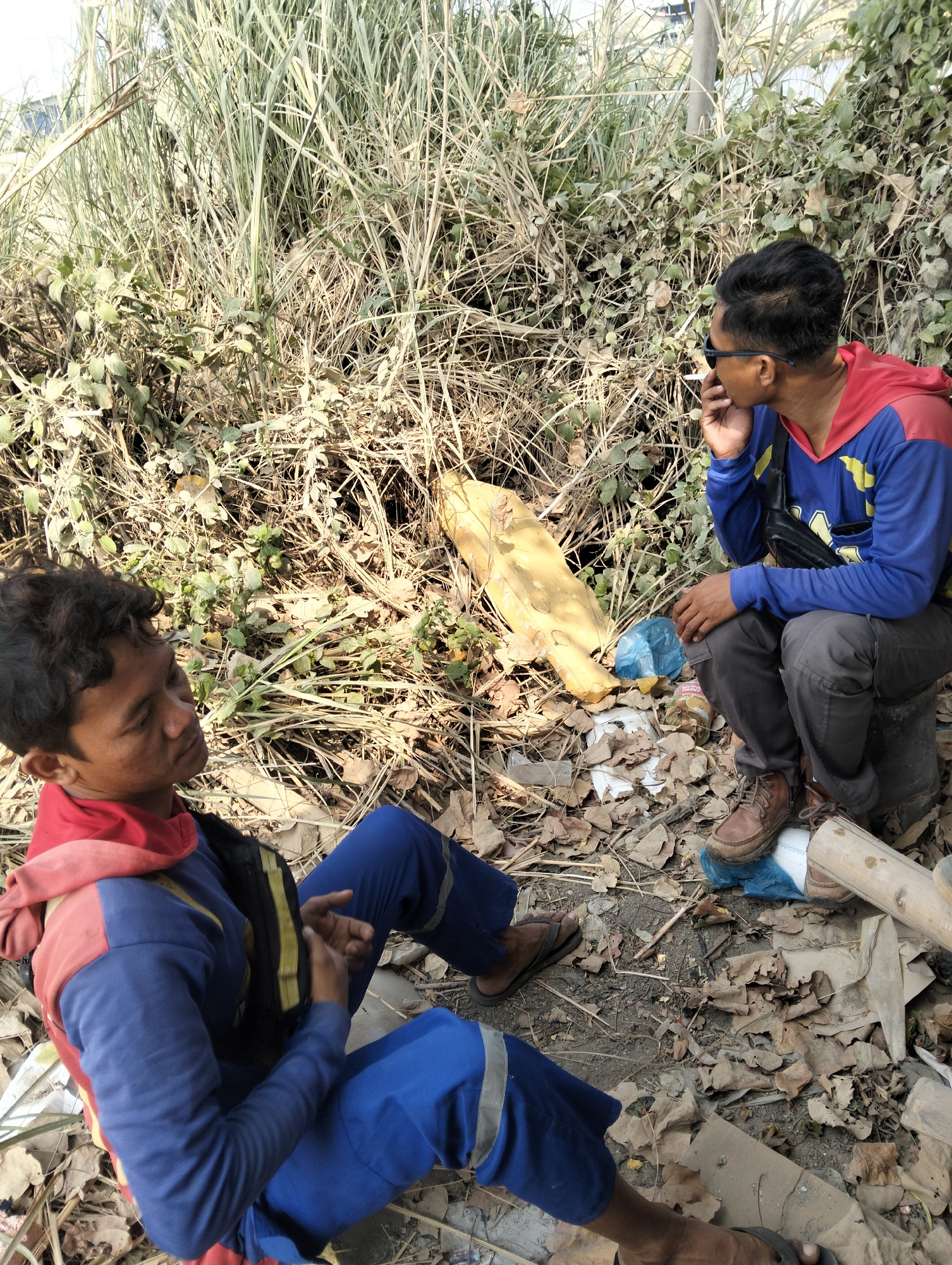 In Semarang, Indonesia, two men are seated on the ground amidst dry foliage and vegetation. The man on the left, wearing a blue jumpsuit with red accents and a yellow harness, is looking down with his hands clasped. The man on the right, dressed in a blue and red hooded jacket, gray pants, and brown shoes, is looking to his left and appears to be smoking. Scattered around them are various objects including a yellow plastic sheet, a blue plastic bag, and a broken piece of bamboo. The environment suggests an outdoor, possibly disused or overgrown area.
