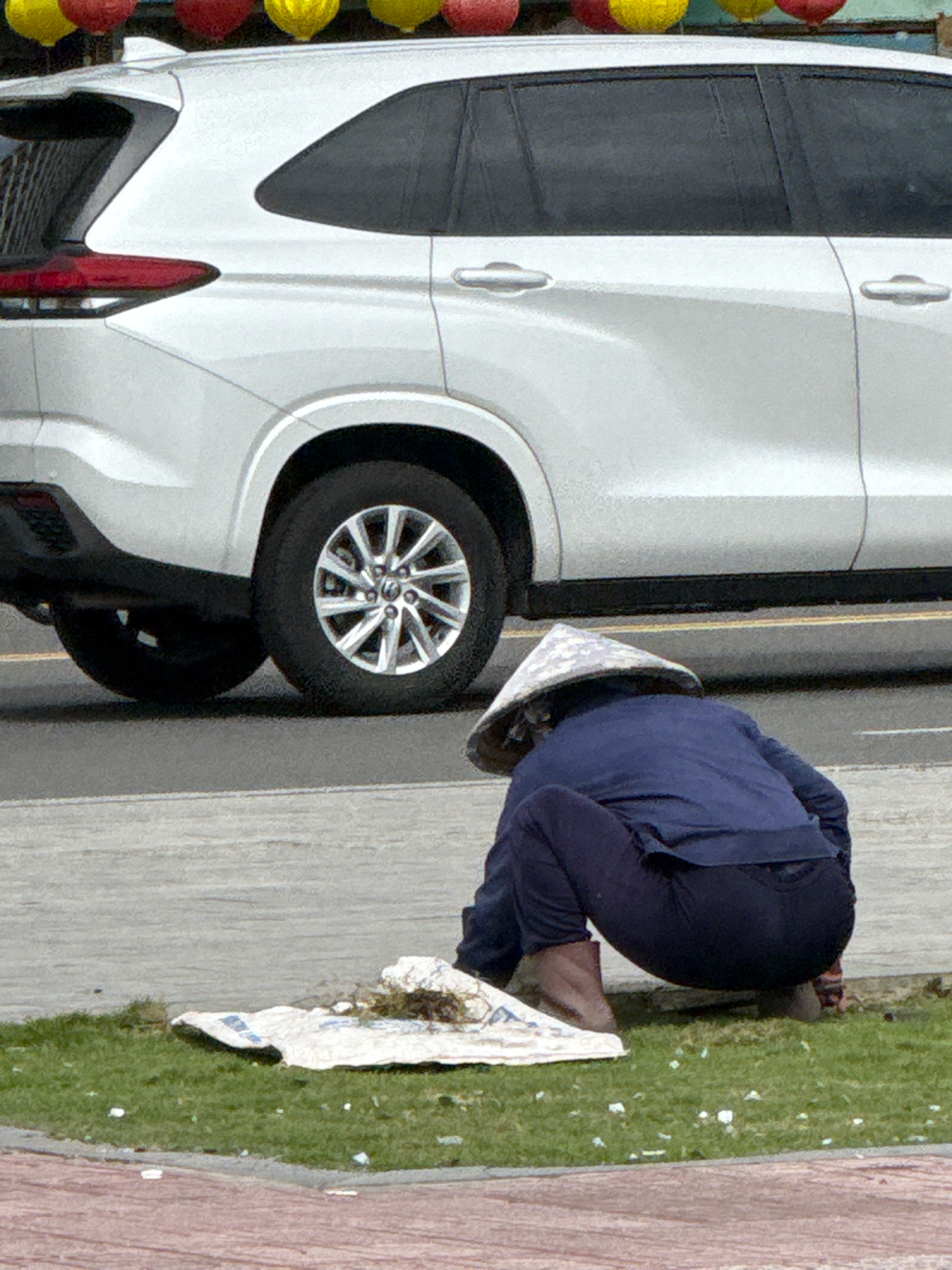 A person wearing a conical hat and a blue jacket is squatting on the grass next to a white SUV in Nha Trang, Vietnam. The person is bent over, possibly cleaning or collecting something from a white cloth laid on the grass. The cloth has some debris on it. The background shows the side of the white SUV, with its tires and part of the road visible. Red and yellow lanterns are hanging in the upper portion of the image. The ground consists of grass, a paved area, and a red brick walkway.