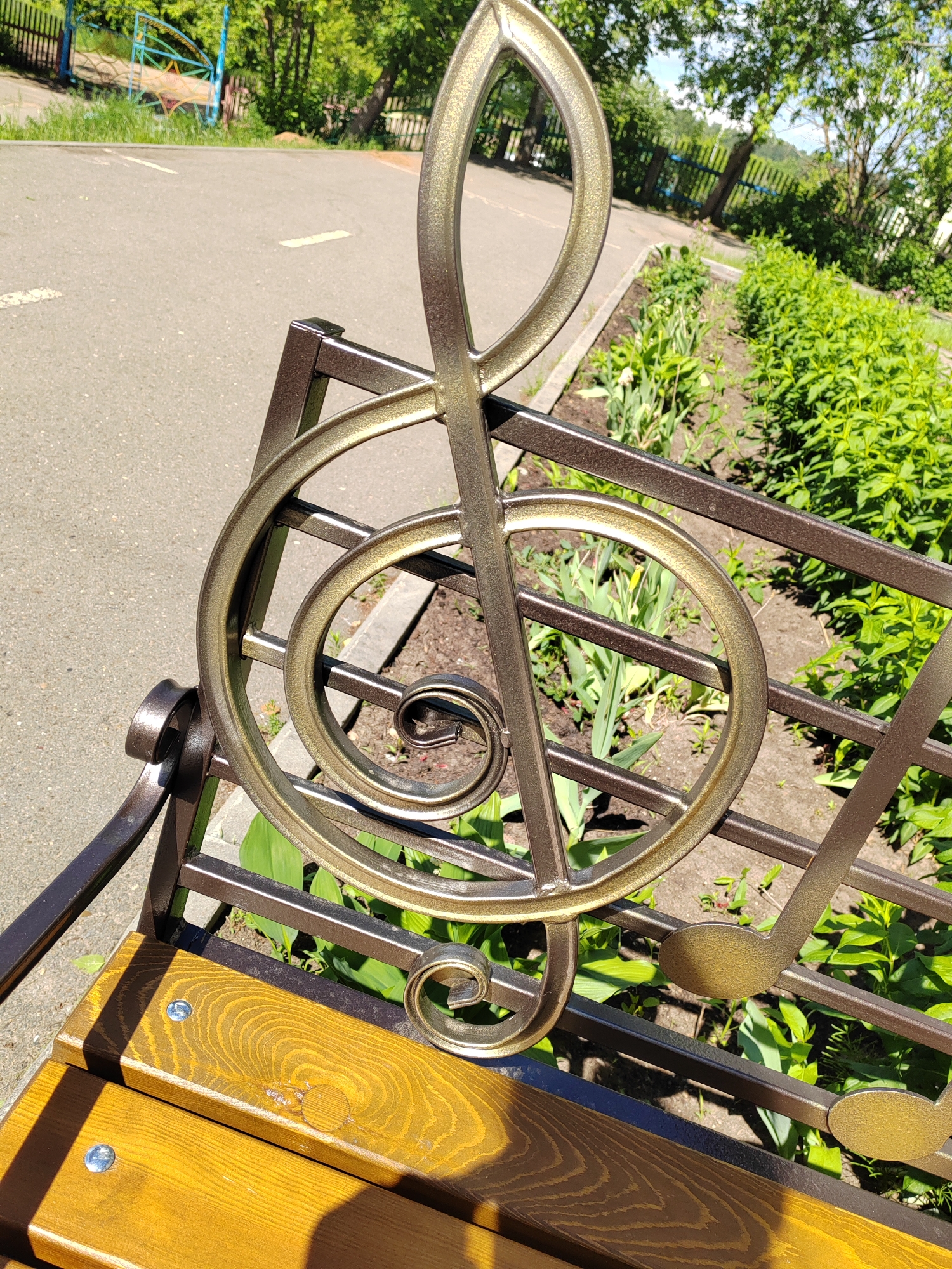 Here is a detailed description of the image:
The image shows a close-up of a park bench with a musical note design incorporated into the backrest. The design includes a prominent treble clef (G clef) made of metal and painted a gold-bronze color. Additional musical notes are also visible in the metalwork.
The bench has a wooden seat made of what appears to be slats, with bolts securing the seat to the metal frame. The frame itself is a dark metal, possibly iron or steel, that complements the gold-bronze of the musical notes.
Behind the bench is a strip of garden bed with green foliage. A paved walkway is visible in the background, along with glimpses of trees and some playground equipment. The scene appears to be outdoors, during the day, in a park-like setting. The location and city are not discernible from the image.