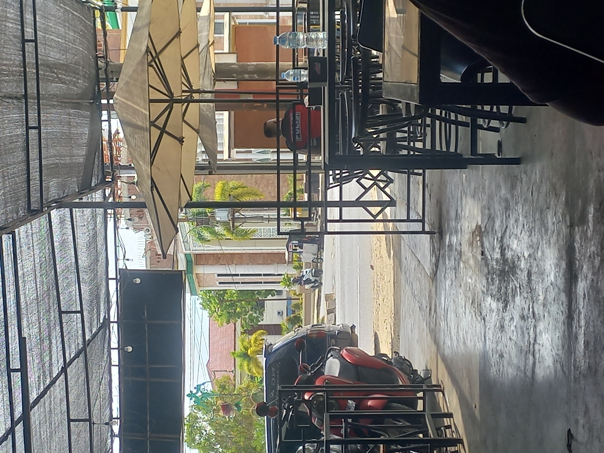 This daytime image captures an outdoor or semi-outdoor cafe or seating area in Titibaro, Indonesia, bathed in bright sunshine. The foreground is dominated by a structure offering shade, featuring a black metal frame supporting a dark mesh tarp roof on the left and several large, light-colored patio umbrellas on the right. Beneath these are multiple black metal tables and chairs, suggesting a casual dining or resting spot.
One person is seated at a table, viewed from behind. They are wearing a red top with the word "POLISI" (Indonesian for "POLICE") clearly visible on the back. Two plastic water bottles are on the table in front of them. The ground beneath the seating area is paved, likely concrete.
Beyond the immediate seating area, a street is visible, with several motorcycles parked or standing, including a prominent red one in the mid-ground. A darker-colored vehicle, possibly a car or van, is also partially visible further down the street. The background features various buildings, likely residential or commercial, characterized by light-colored walls and tiled roofs, interspersed with lush green foliage, including palm trees. The clear sky indicates fine weather. The scene portrays a typical, relaxed street-side environment.
