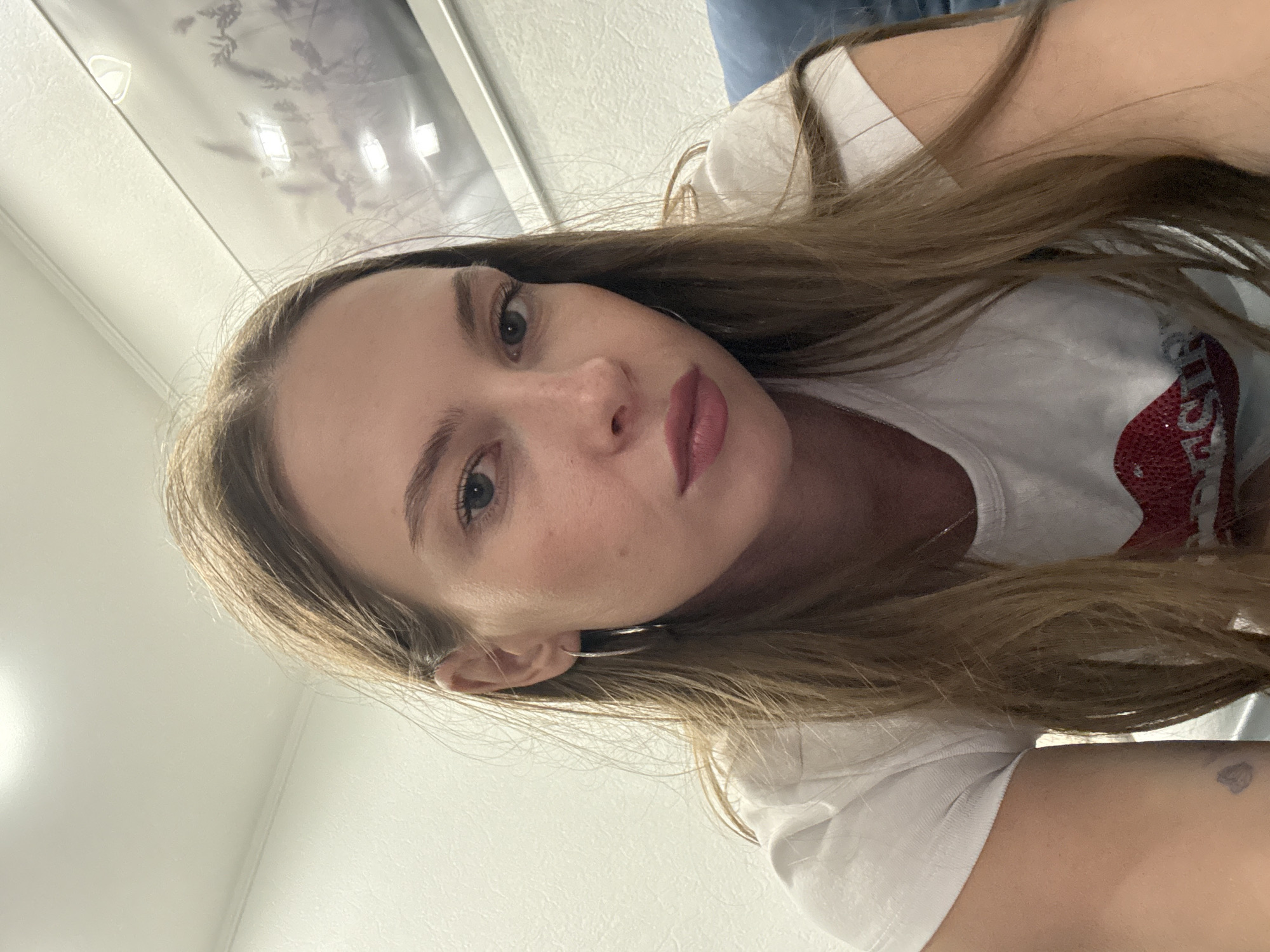 The media file is a selfie of a young woman lying down. She has long, light brown hair that falls around her face and shoulders. She is wearing a white t-shirt with a red glittery design on it, possibly a heart or lips. The woman has light skin, green eyes, and is wearing lipstick. She has a subtle smile and appears relaxed. There's a faint shadow on her arm. The background is a plain white wall and a glimpse of a light-colored ceiling with some texture. The lighting is soft and diffused, suggesting it could be daytime indoors. No specific location cues for Perm, Russia are visible in the image itself. The overall mood is casual and personal.