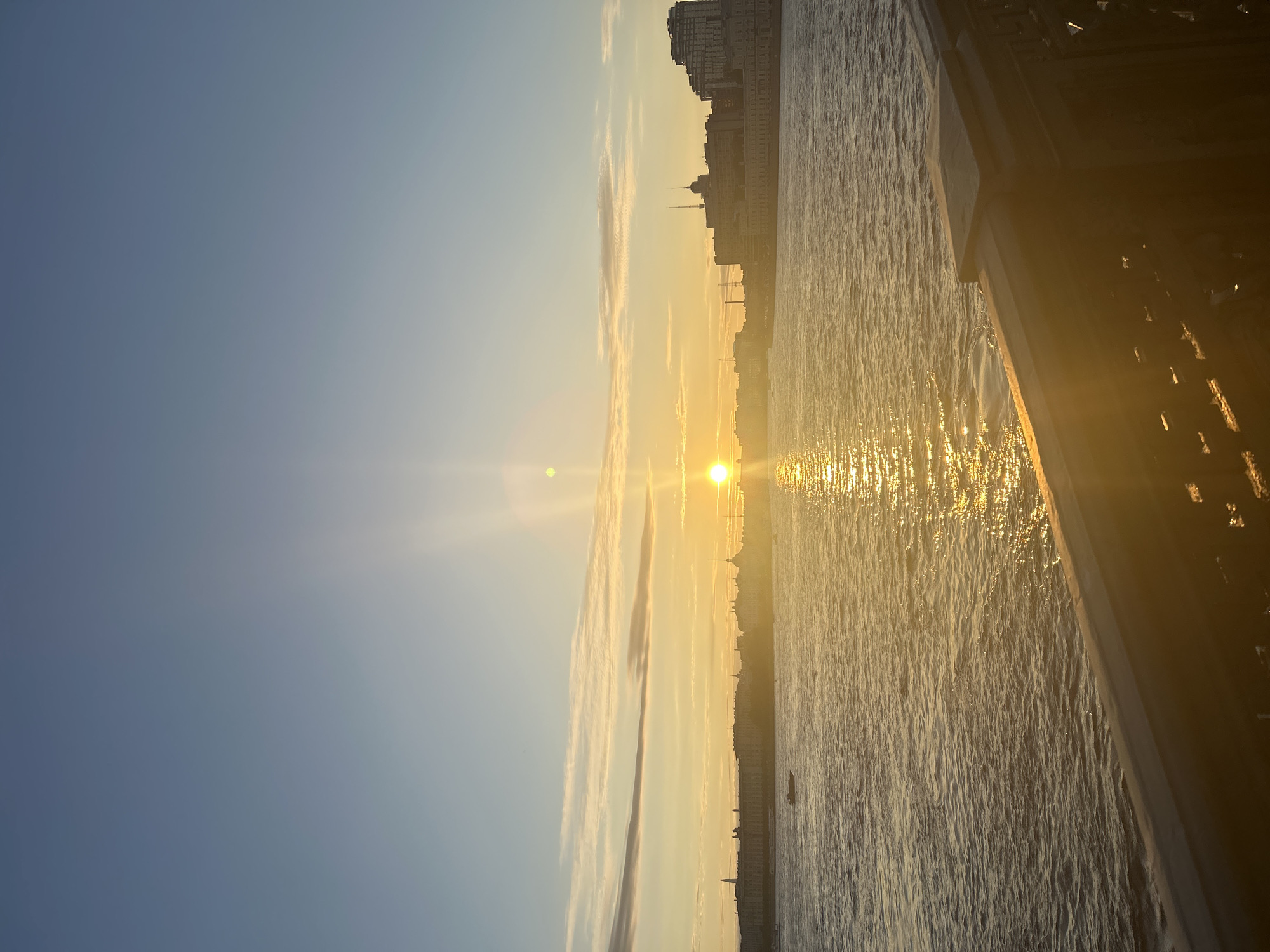 A vertical, eye-level shot captures a sunset over the Neva River in Saint Petersburg, Russia. The sun, a bright orb of light, sits low on the horizon, casting a golden glow across the water and the sky. A distant cityscape lines the riverbank, with buildings of varying heights and architectural styles silhouetted against the colorful sky. The water is calm, with gentle ripples reflecting the sunlight. A small boat navigates the river, its silhouette adding a touch of movement to the scene. In the foreground, the dark, ornate railing of a bridge or embankment frames the view, its metallic texture catching the warm light of the sunset. The sky transitions from a clear blue at the top to a soft yellow and orange near the horizon, with a few wispy clouds adding texture.