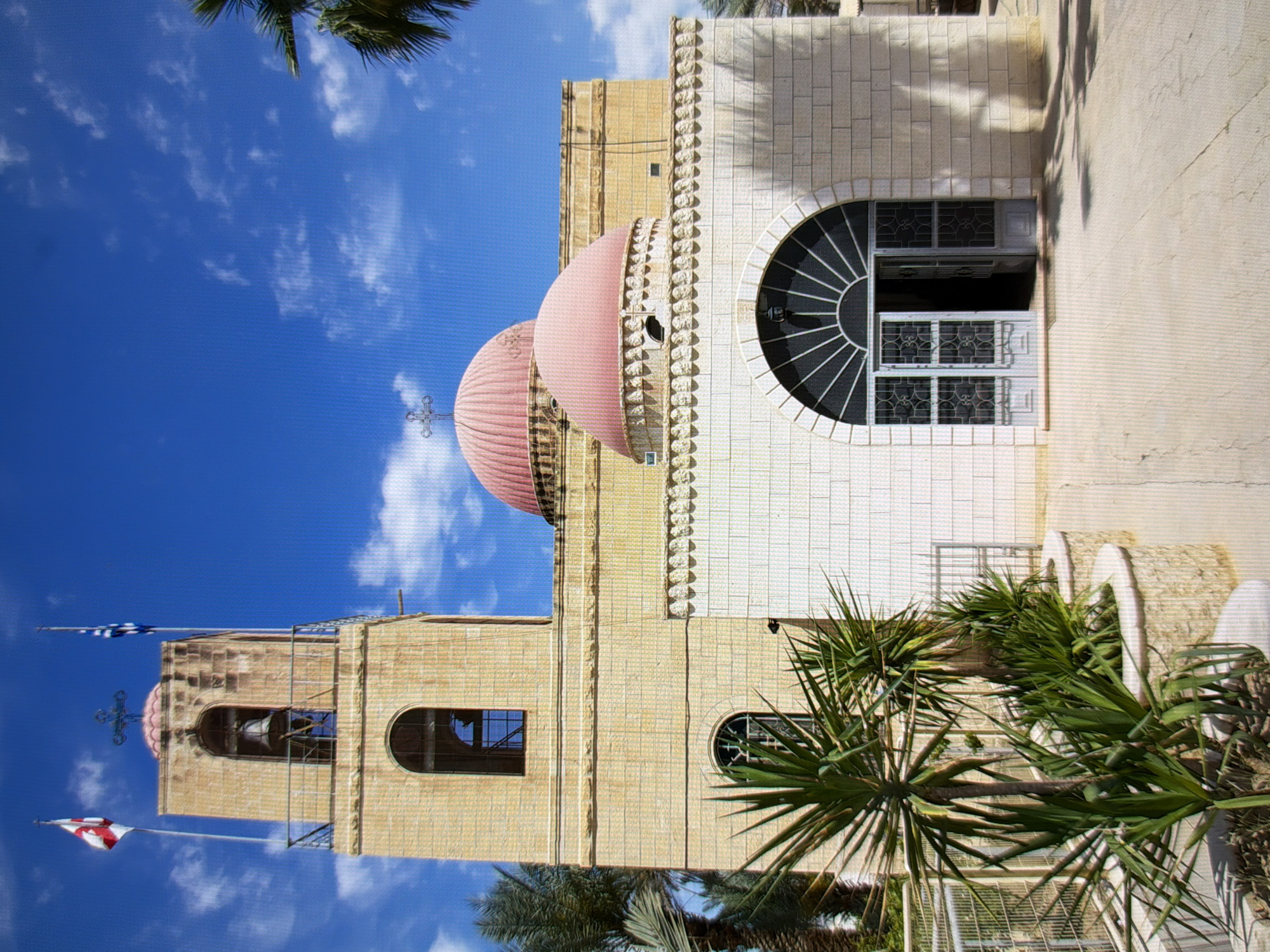 This media file depicts a large, light-colored stone building, possibly a church or a significant institutional structure, under a bright blue sky with scattered white clouds. The building features architectural elements such as domes, arched windows, and decorative stone friezes. Two flags are visible: one is a blue and white checkered flag, likely representing Greece, and the other is a red and white striped flag, which could be the flag of Saint George or a local variation. A cross is visible atop one of the domes. Lush green palm trees and other foliage are present in the foreground, suggesting a warm climate. The image is taken from a low angle, looking upwards towards the building. The lighting suggests it is daytime, likely mid-morning or afternoon, with bright sunlight casting shadows. No people are visible in the frame. The image's orientation is rotated, making it appear sideways. While the location context provided is Kyiv, Ukraine, the visual cues such as palm trees and the architectural style do not strongly align with typical Ukrainian landscapes and architecture, hinting at a possible discrepancy or a specific, less common location within Kyiv.