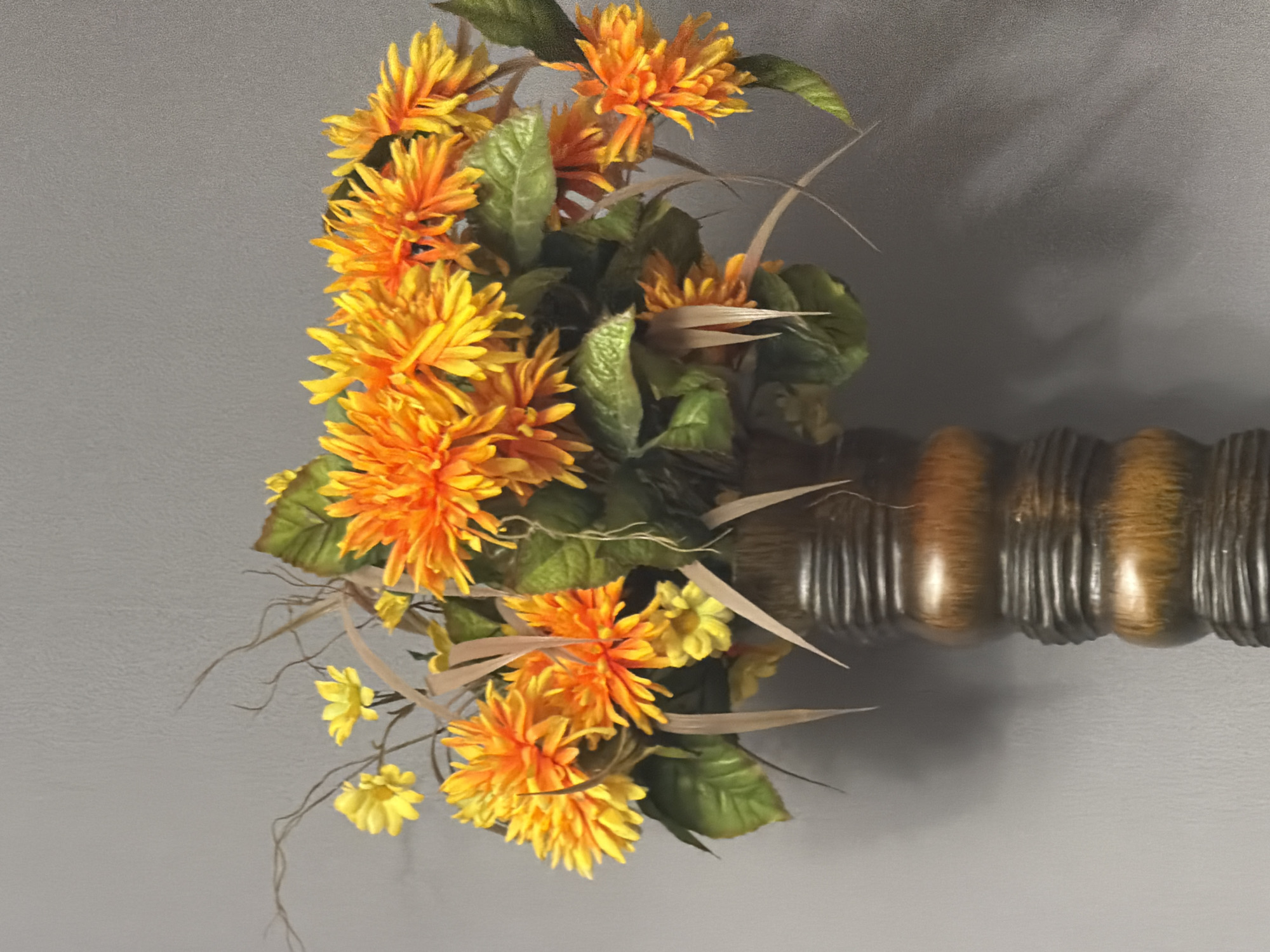 A floral arrangement of bright orange and yellow artificial dahlias and small yellow flowers is displayed in a tall, dark brown, ribbed vase. The vase has a glossy finish and a segmented, bulbous shape. The arrangement includes green artificial leaves and wispy, light brown decorative elements that resemble dried grass or twigs. The background is a plain, textured gray wall, against which shadows from the arrangement are cast. The image is taken from a slightly low angle, looking up at the floral display.