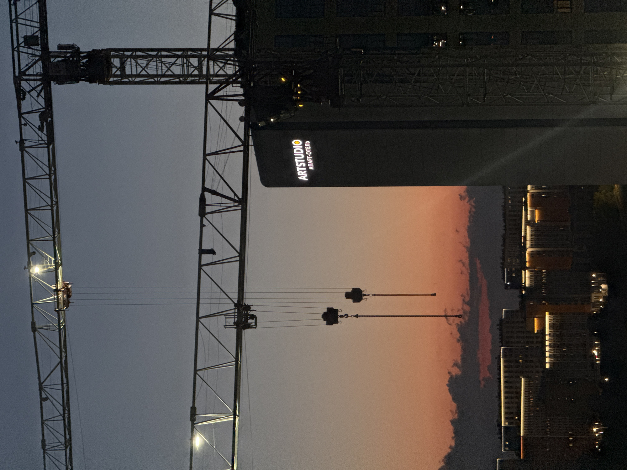 The image depicts a construction site at dusk in Sinyavino, Russia. Two tower cranes dominate the foreground, with their booms extending horizontally across the frame. The sky displays a gradient of dark blue at the top to a warm orange and pink near the horizon, indicating twilight. In the mid-ground, a building is illuminated with the sign "ARTSTUDIO АПАРТ-ОТЕЛЬ" displayed prominently on its side. Below this, a cityscape of apartment buildings is visible, featuring illuminated windows and decorative lighting along their rooftops. The lower portion of the image shows the dark silhouettes of trees or other vegetation. There are no visible people or active construction activities in this static scene.