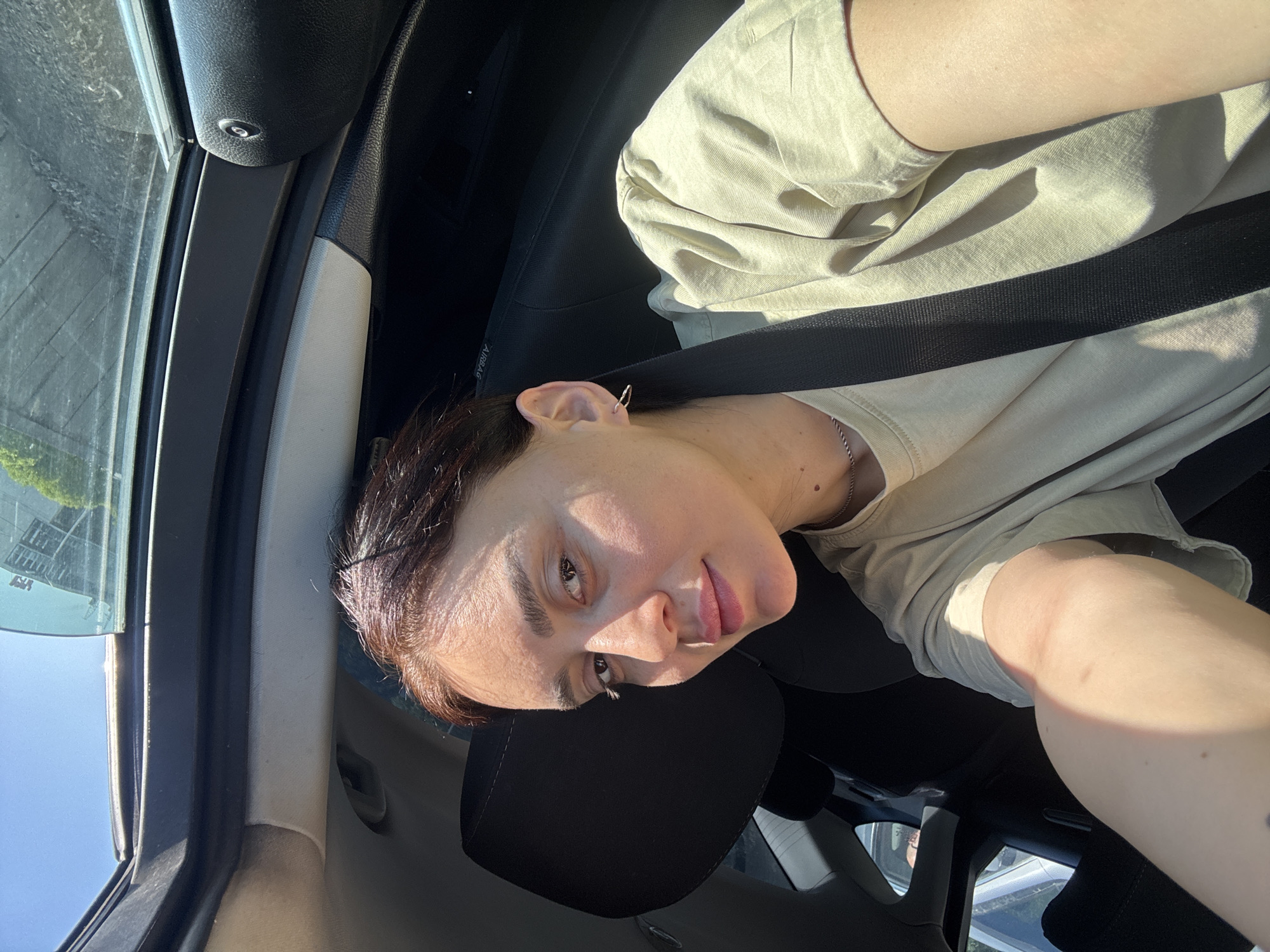 A person is in the driver's seat of a vehicle, wearing a beige t-shirt, and a seatbelt. The image appears to be taken from inside the vehicle, with the front window and interior visible. The surrounding environment outside the vehicle includes buildings and other vehicles. The location of the city of Tbilisi, Georgia is not evident in the picture.