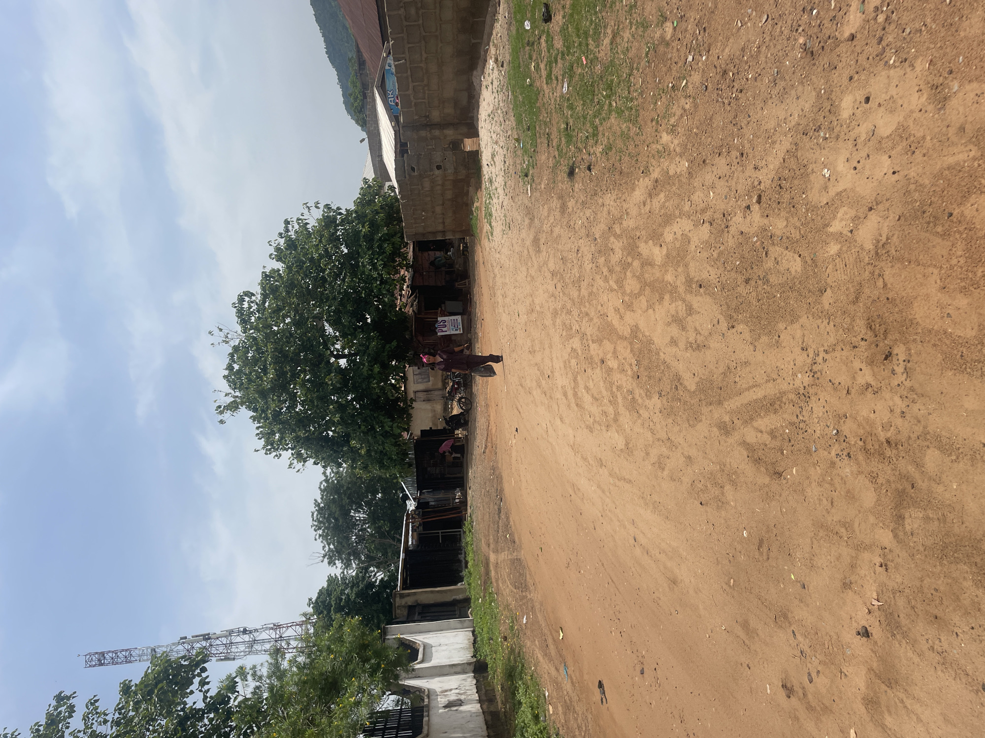 The image depicts a scene on a dirt road in Lokoja, Nigeria. A person, dressed in dark clothing and a headscarf, walks away from the camera, carrying a dark bag. The road is unpaved and appears to be a dirt track, with tire marks visible. On either side of the road, there are various structures, including a white-walled building on the left, some makeshift stalls or shops with corrugated metal roofs, and a brick wall on the right. A cell tower is visible in the distance, behind trees. The sky is overcast with some clouds. The overall impression is a semi-urban or rural environment.