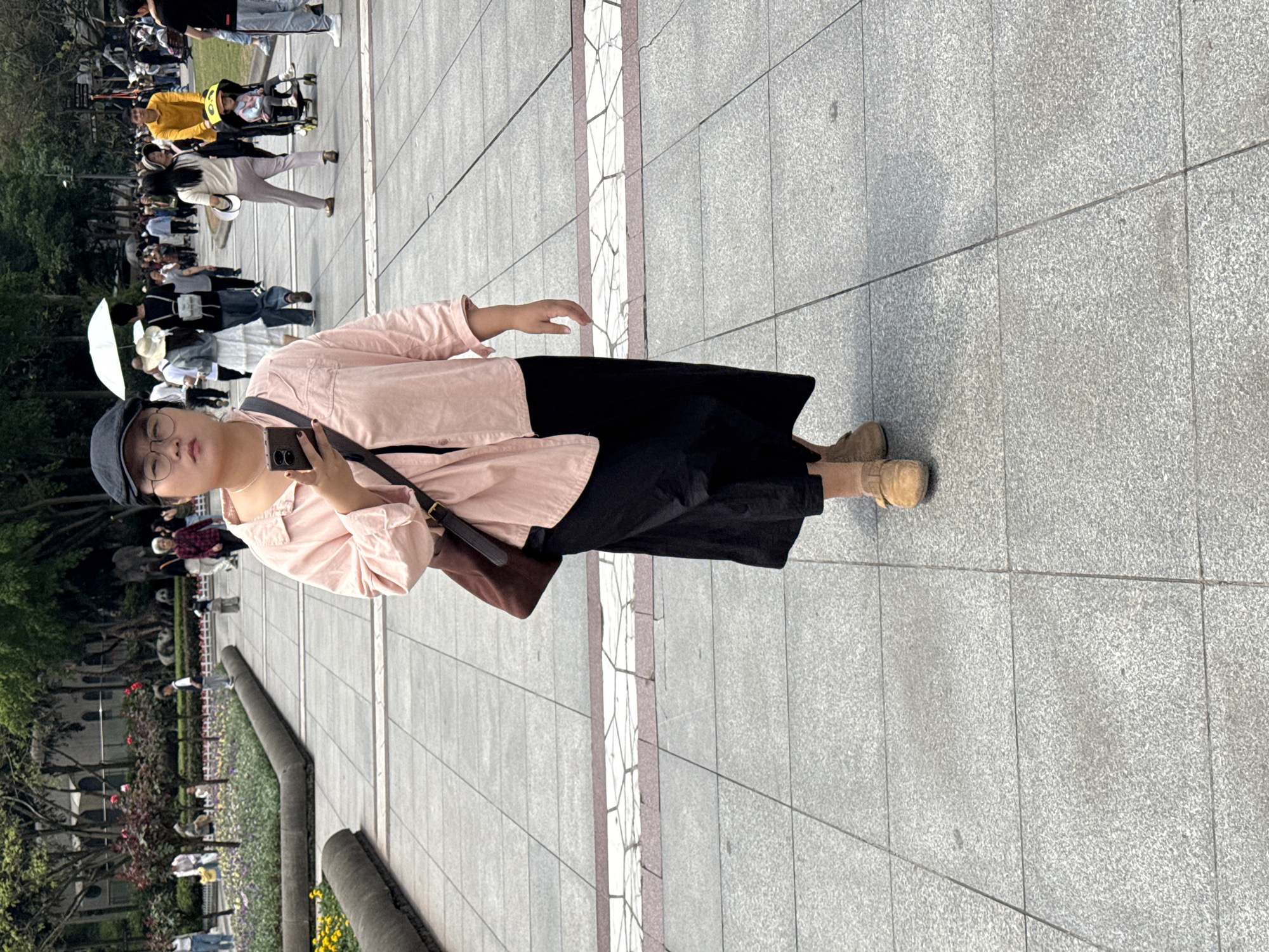 A person wearing a pink shirt, black pants, a cap, and glasses stands on a paved area while holding a phone. The person is looking at the phone. In the background, there are other people, trees, flowers, and low walls. The scene appears to be a public park in Jiulongpo District, China.