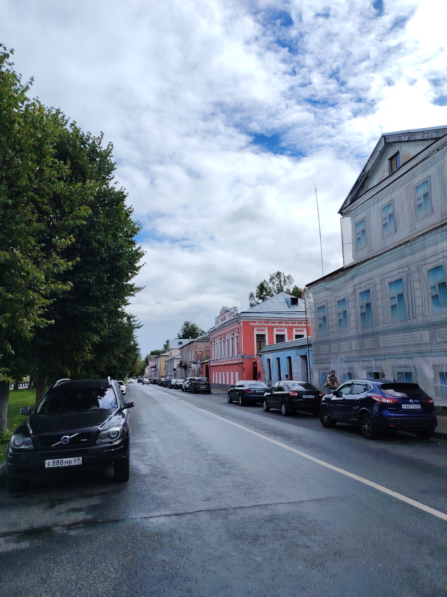 The image captures a street scene in Tver, Russia, featuring a row of historic buildings lining the right side of a wet asphalt road. A large, mature tree with dense green foliage dominates the left side of the frame. Several cars are parked along the street and a few are in motion, including a black Volvo SUV in the foreground on the left and a blue Nissan SUV on the right. The buildings exhibit varied architectural styles and colors, with some featuring trompe-l'œil murals that create the illusion of detailed facades. The sky above is partially cloudy with patches of blue visible. The overall impression is a quiet, slightly overcast day in a European city, with a blend of old-world charm and modern transportation.