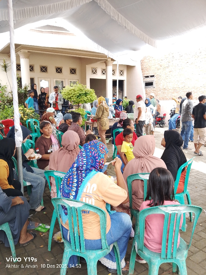 A gathering of people sits on green plastic chairs under a white tent. Some are eating from plates. A two-story building with decorative windows is visible in the background, as are some trees and shrubbery. Some people are standing and mingling, while others are seated. A parked motorcycle can be seen in the distance.