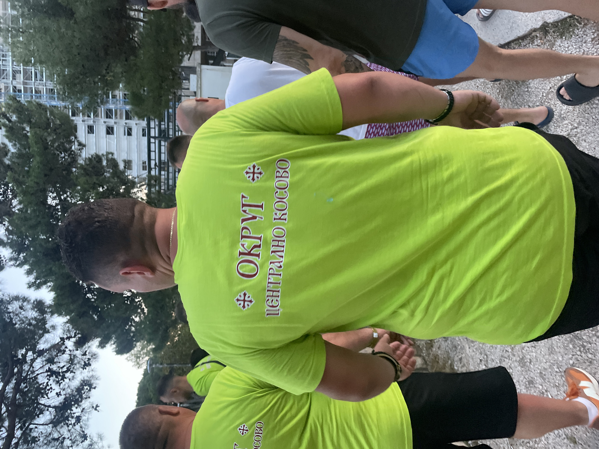 The image shows a group of men from behind, wearing neon green t-shirts. The t-shirts have Cyrillic writing on them. The man in the foreground has "ОКРУГ ЦЕНТРАЛНО КОСОВО" written on his shirt, which translates to "District Central Kosovo." Another man to the left has "осово" visible on his shirt. The men are standing on a gravel path, with trees and a tall building under construction in the background. The lighting suggests it is daytime, possibly late afternoon.