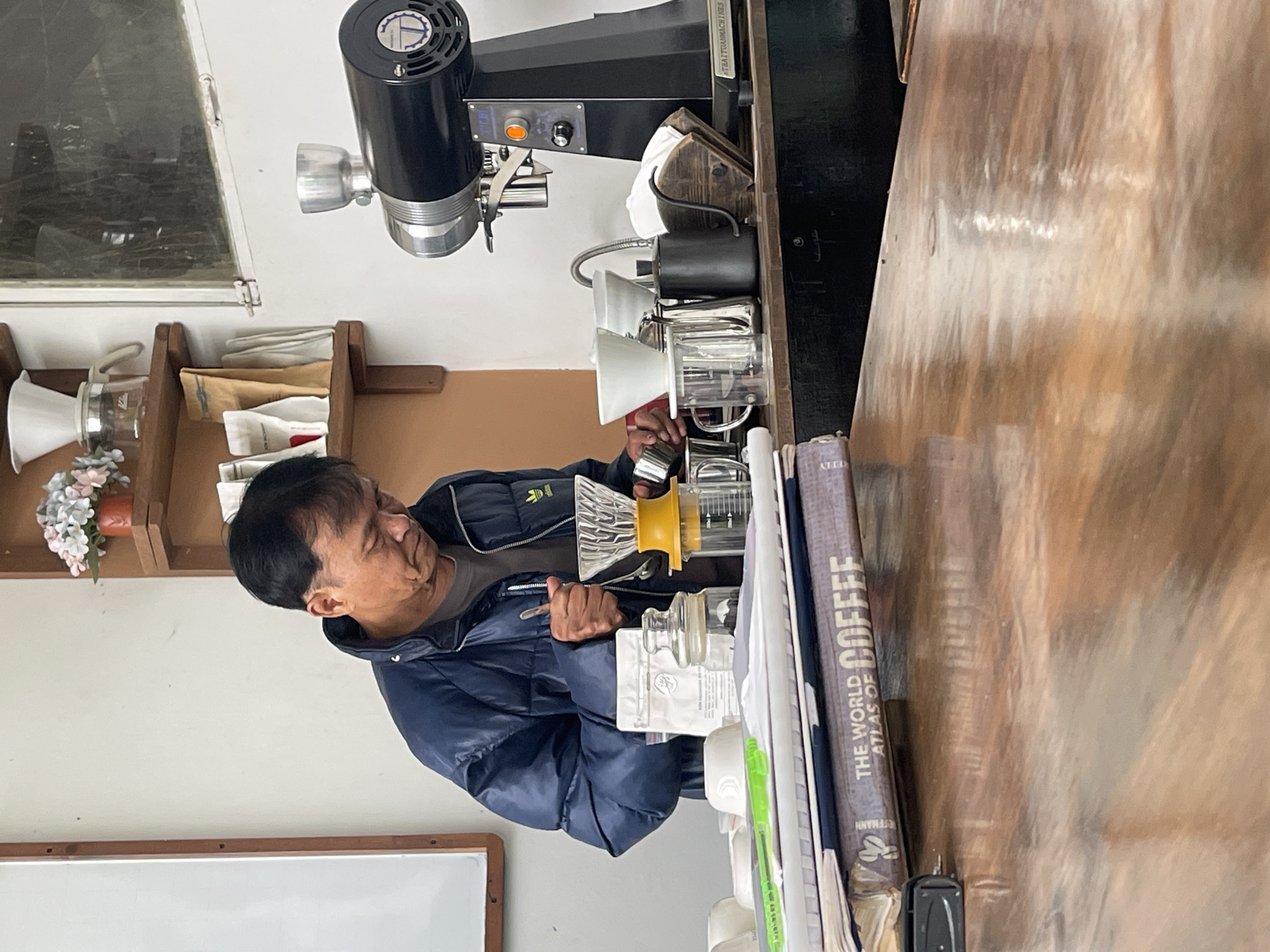 In Cam Ly - Da Lat, Vietnam, a man wearing a navy-blue puffer jacket is shown preparing coffee at a counter. He is holding a yellow-ringed glass coffee dripper in his hands, with additional glass carafes and a white dripper nearby. A coffee roaster machine is partially visible above and behind the drippers. On the right side of the image, a stack of books including "The World Atlas of Coffee" rests on the counter. Behind the man is a shelf holding items including coffee filters, and above the shelf is a window. To the bottom left of the man is a whiteboard.